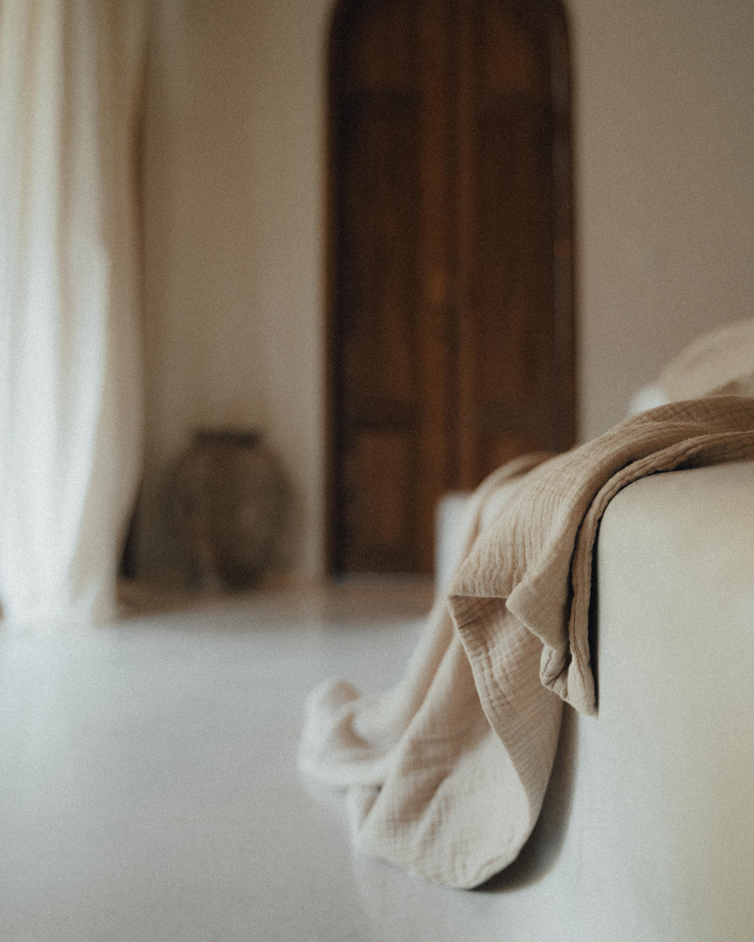 Minimalist bedroom with a soft beige blanket on the bed, a curtain, pottery, and a wooden door in the background.