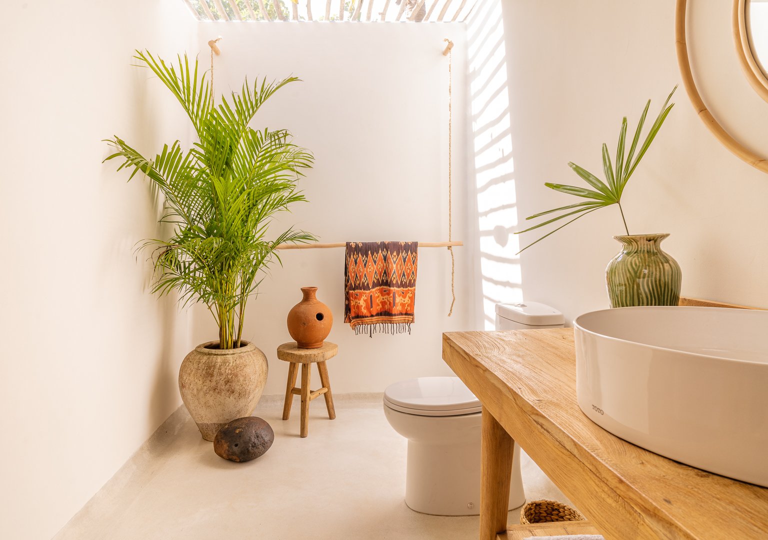 Minimalist bathroom with natural accents, including a large potted plant, wooden stool, ceramic vase, and a woven basket. The room features a modern toilet, wooden countertop with a round white basin, and a colorful towel hanging on a wooden bar.