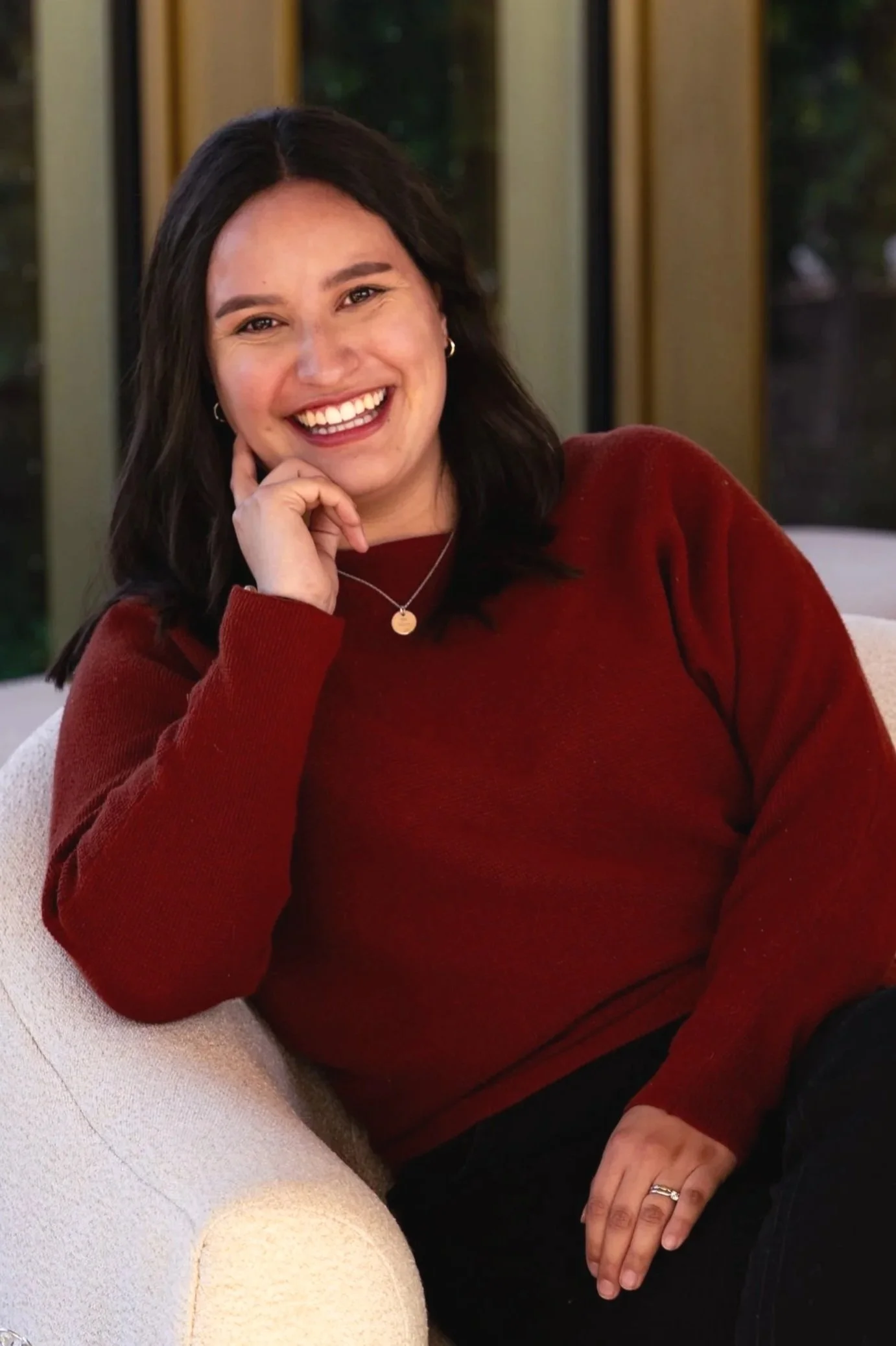 Young woman with dark hair, smiling and wearing a red sweater, seated on a light-colored chair with a glass window and greenery in the background.