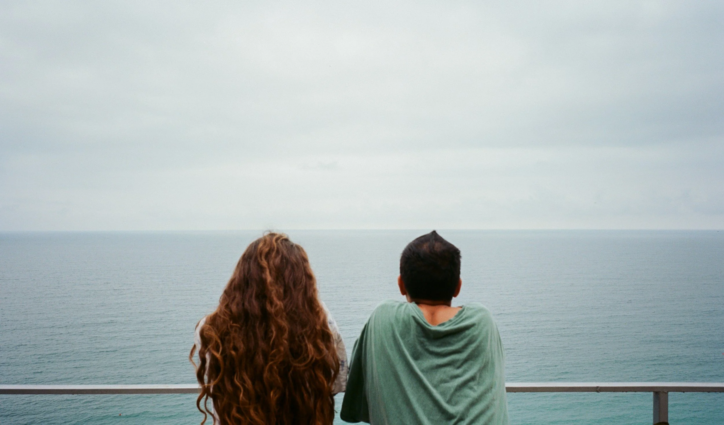 A woman and a man with their backs to the camera, sitting on a railing and looking out over the ocean on a cloudy day.
