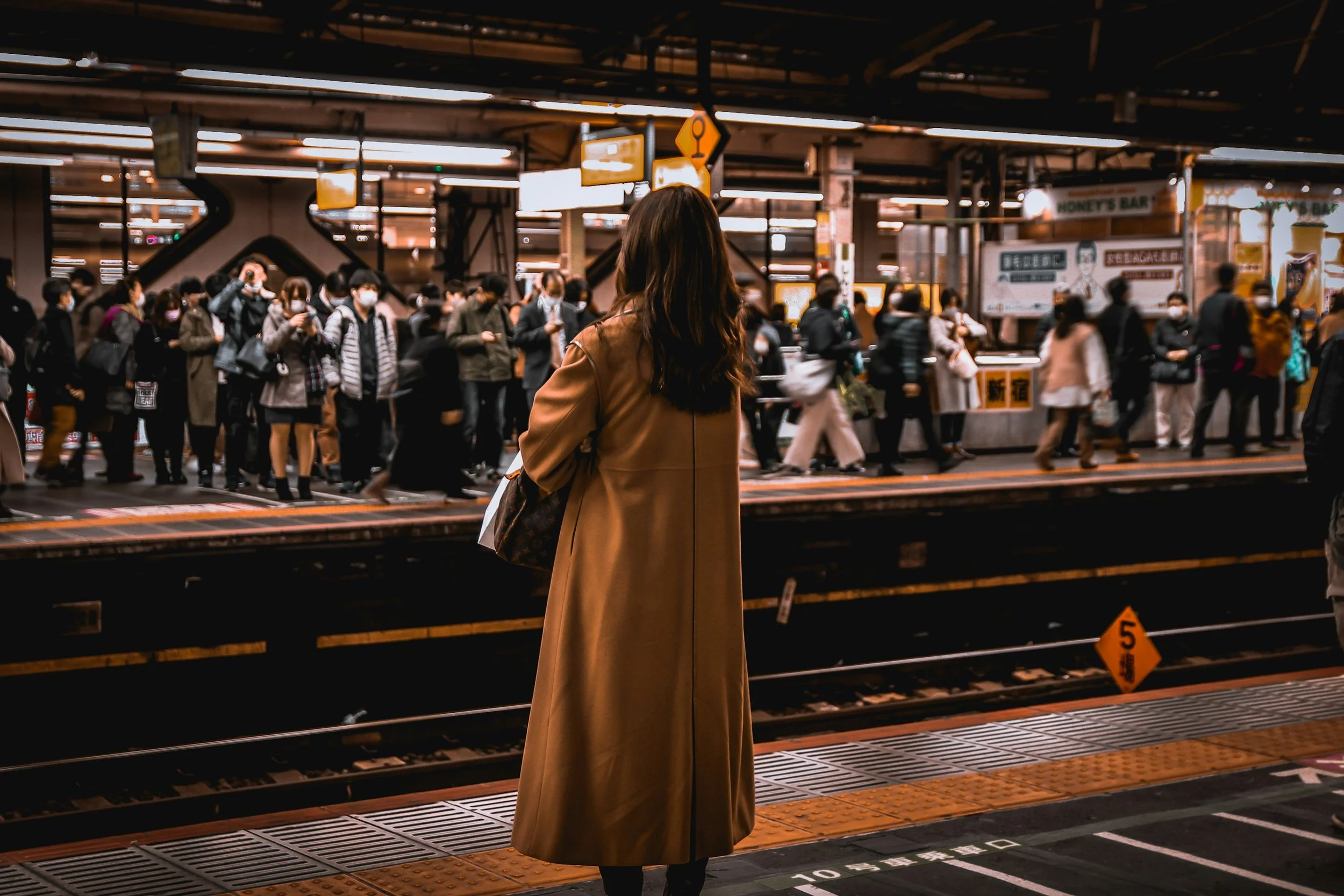 A woman standing alone on a subway platform looking toward the tracks, with people moving in different directions on the opposite side