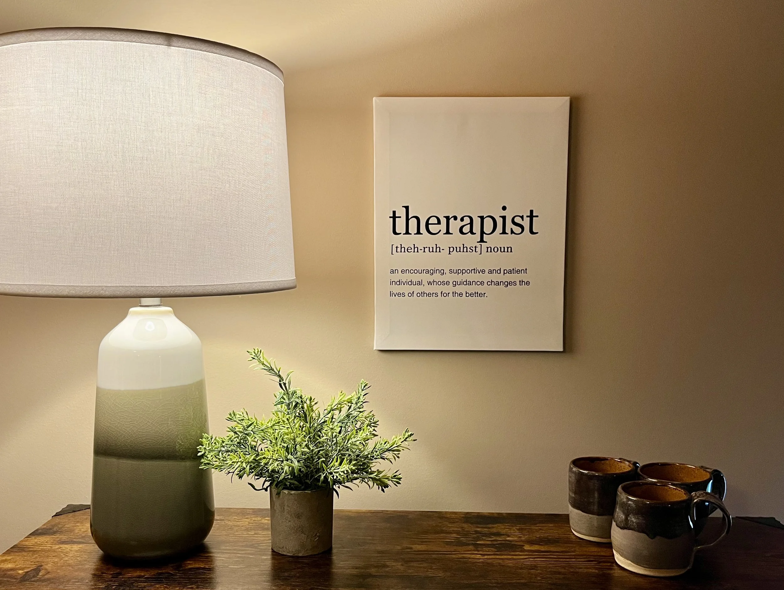 Cozy room corner with a table lamp, potted plant, wall art, and three mugs on a wooden surface. The wall art features a definition of 'therapist.'
