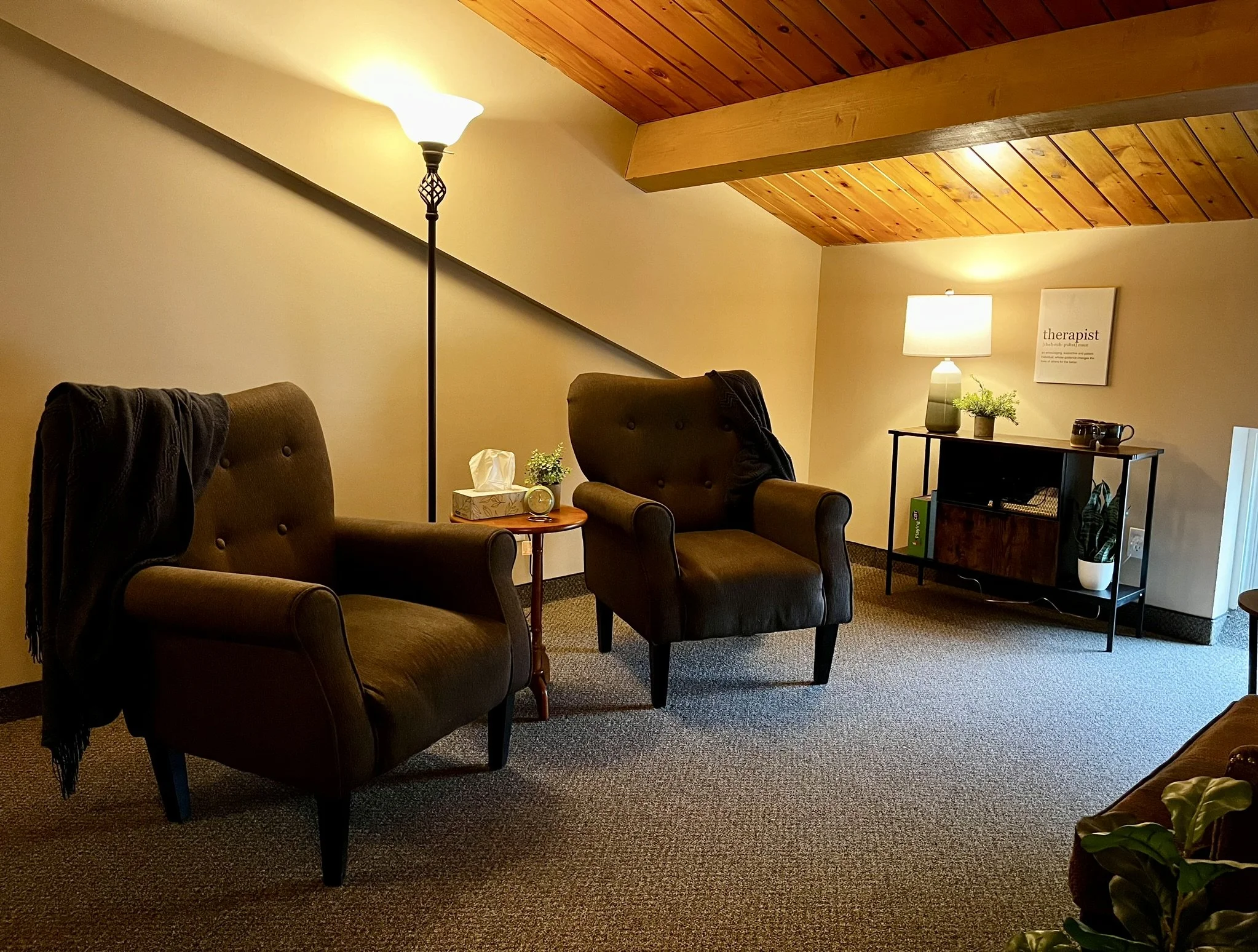 Cozy therapy room with two upholstered armchairs, a side table, tissues, and soft lighting. A floor lamp and a table lamp provide ambient light. Decor includes plants and a small shelf unit with a mug and a book. Sloped wooden ceiling and carpeted floor enhance warmth.