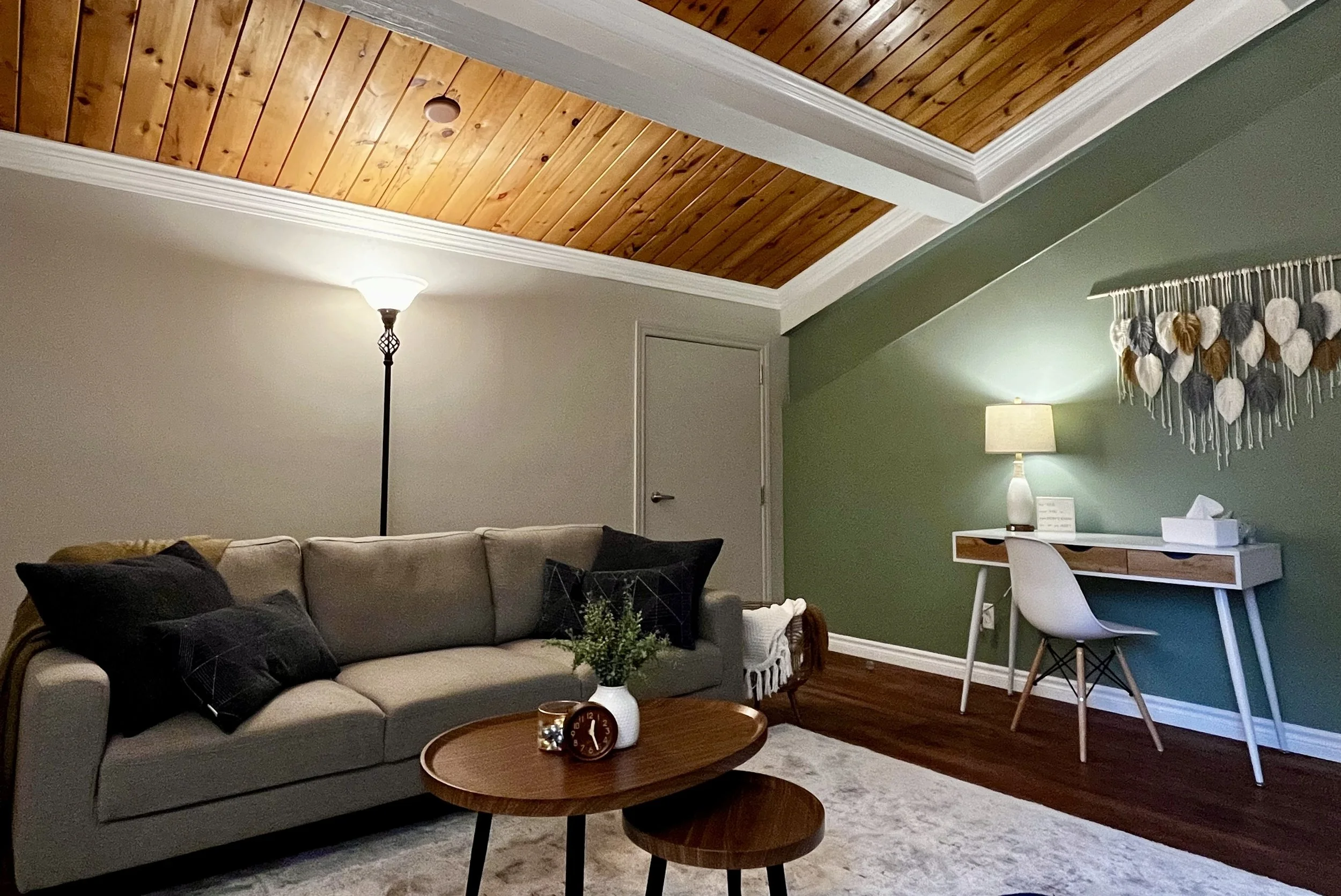 Stylish living room with wooden ceiling, gray sofa, floor lamp, round coffee tables, desk with chair and lamp, and wall decor on green accent wall.
