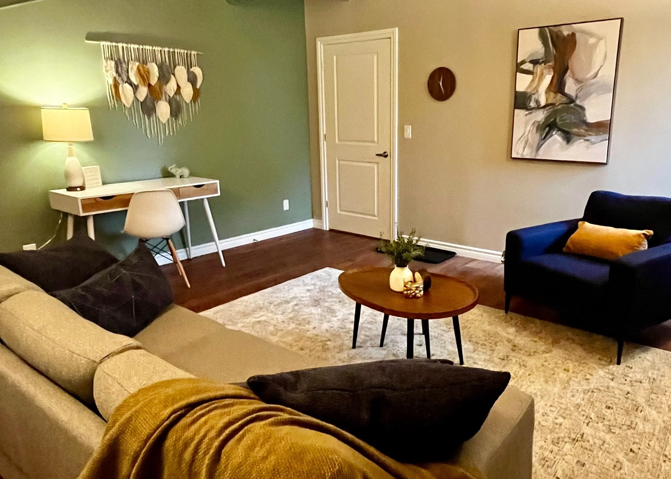 Cozy living room with a green accent wall, white desk, chair, and decorative weaving. A modern painting hangs on a beige wall, and there's a small wooden coffee table with a vase and decor. A blue armchair and beige couch with a throw blanket are also visible.