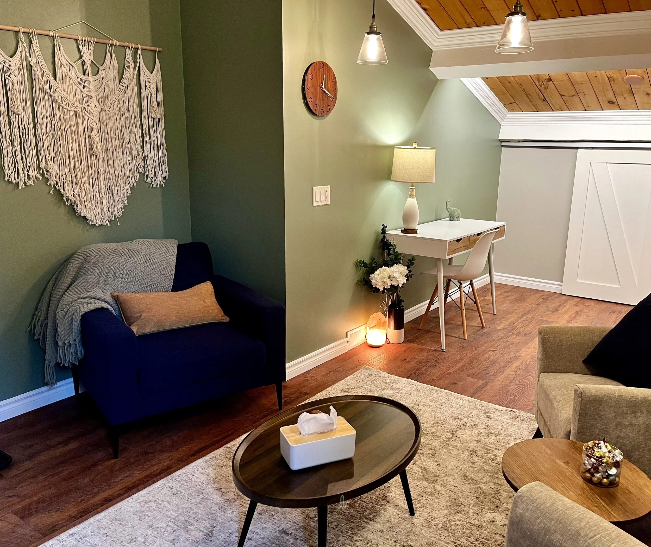 Cozy living room with green walls, featuring a dark blue armchair with a pillow and throw blanket, a wall-hanging macramé decoration, a small round wooden coffee table with tissues, a white writing desk with a chair and lamp, decorative flowers, a wall clock, and warm lighting from pendant and table lamps.