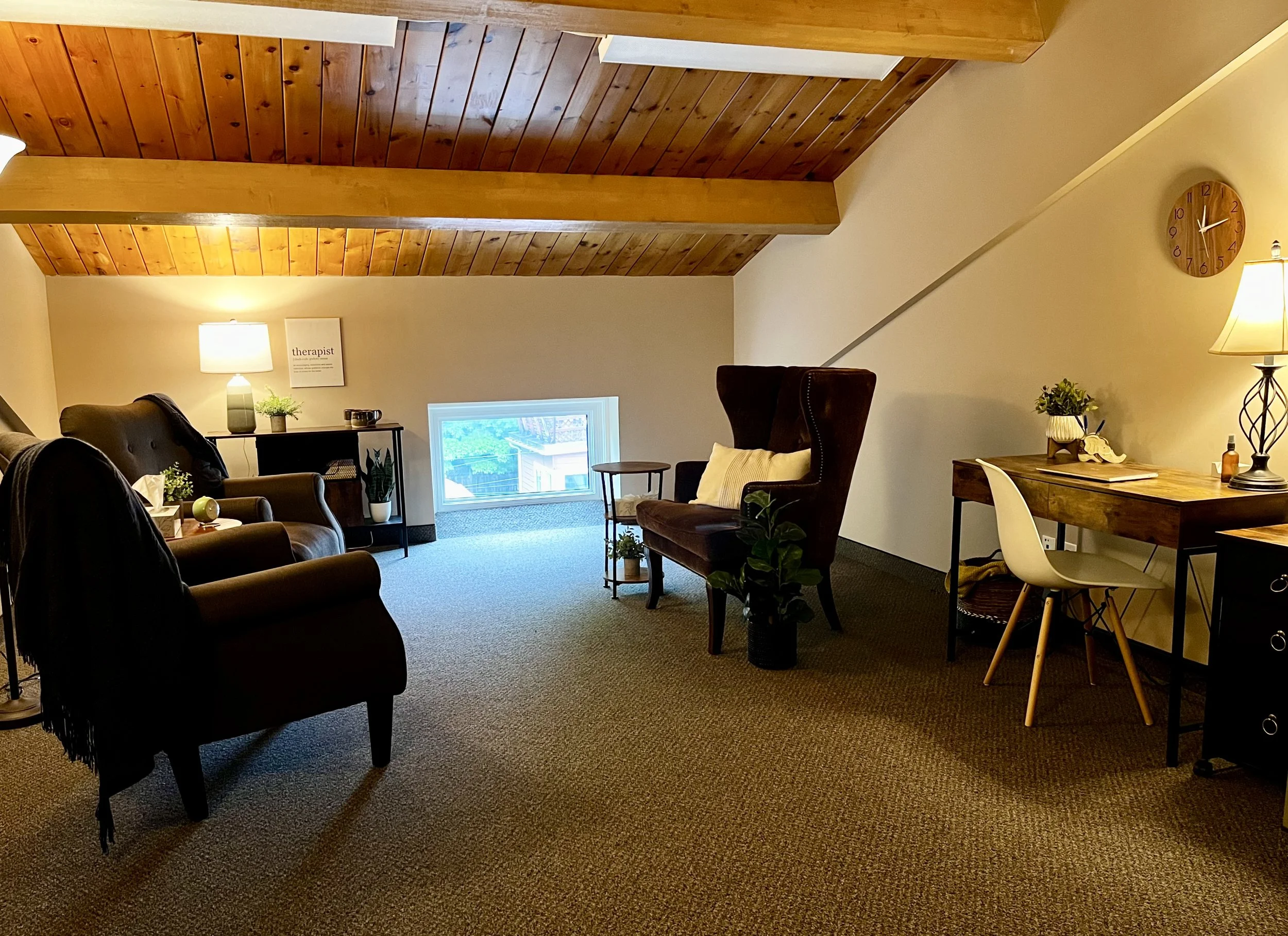 A cozy attic office with wooden ceiling, armchairs, a wooden desk with a lamp, indoor plants, and a small window.