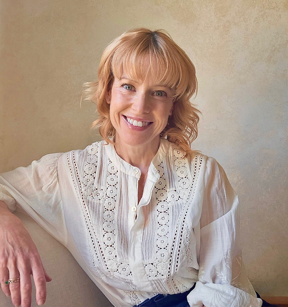 A woman with shoulder-length blonde hair wearing a white eyelet blouse, smiling and sitting on a beige couch against a plain beige wall.