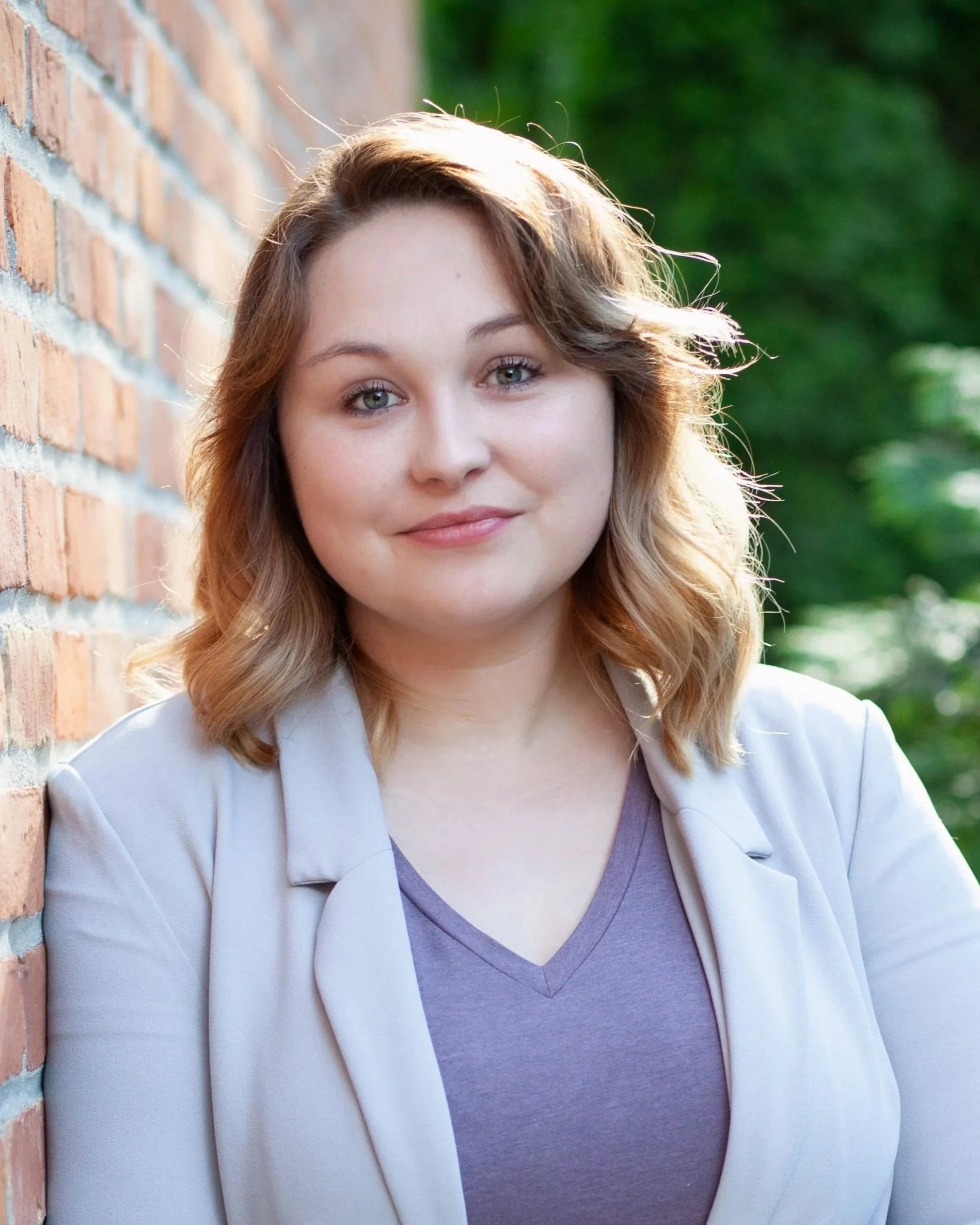 A young woman with shoulder-length wavy hair, standing outdoors next to a brick wall, with green foliage in the background, wearing a light-colored blazer and a purple V-neck shirt, smiling softly at the camera.