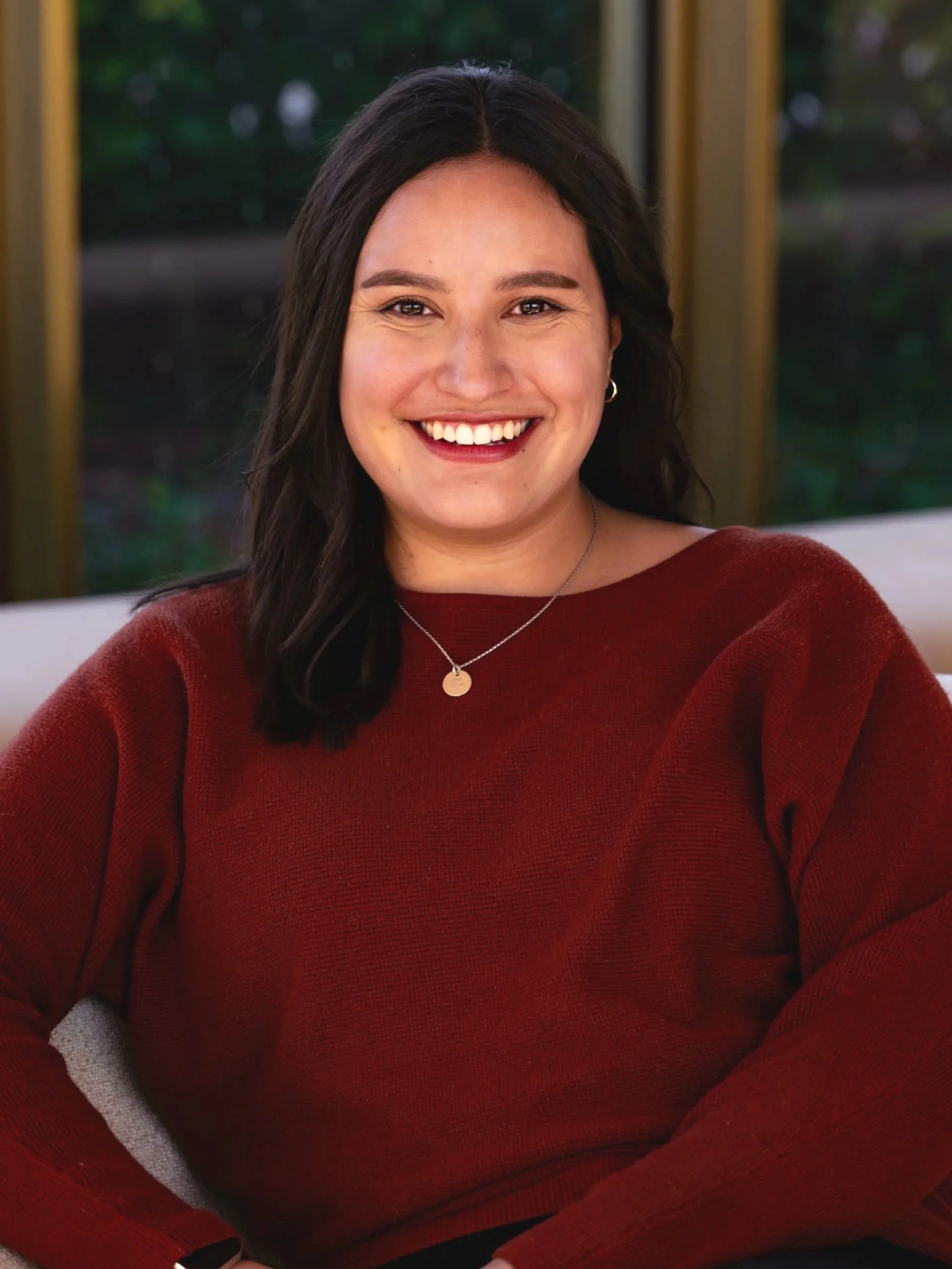 Young woman with dark hair, smiling and wearing a red sweater, seated on a light-colored chair with a glass window and greenery in the background.