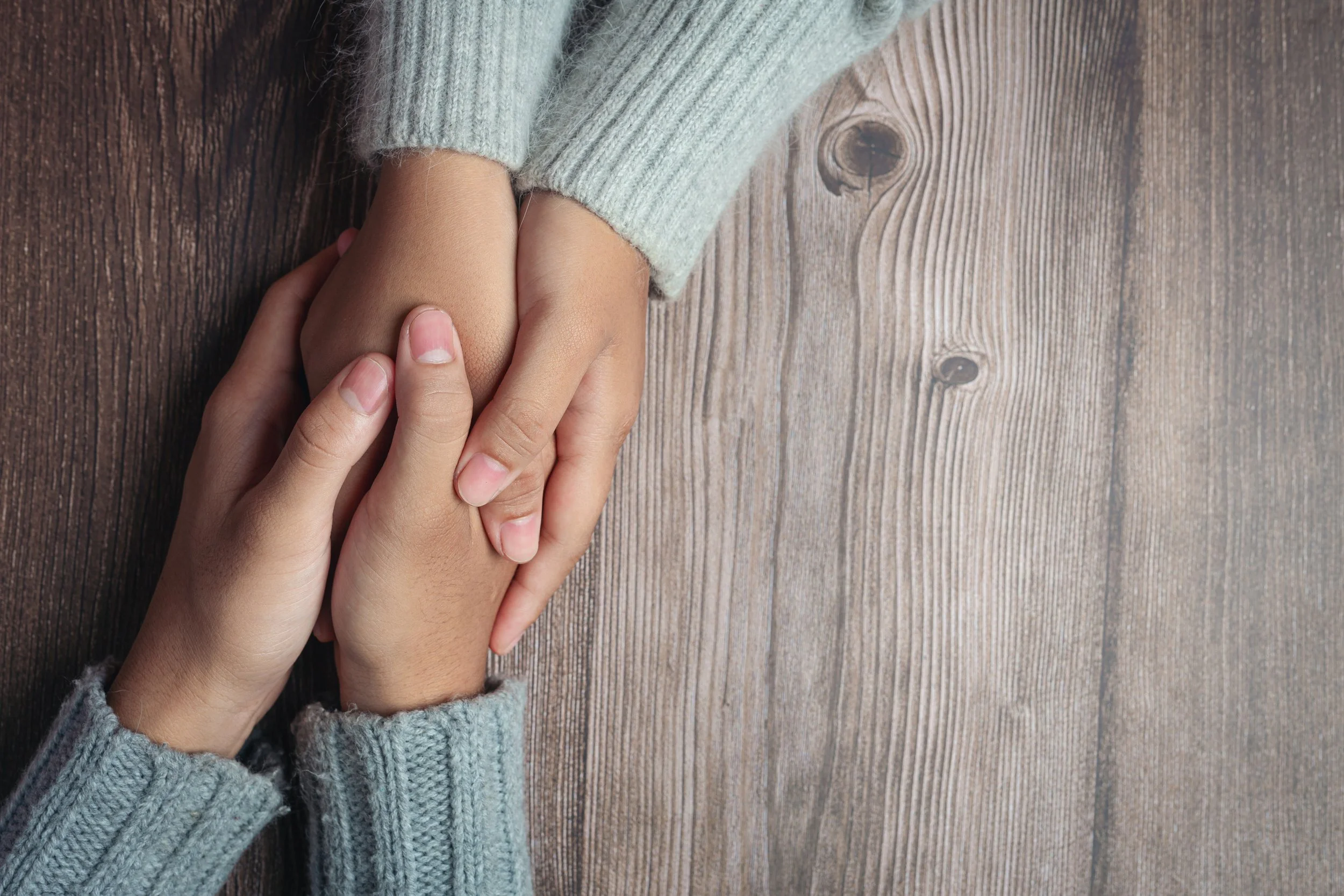Overhead image of two people clutching each other's hands supportively against a wooden table surface.