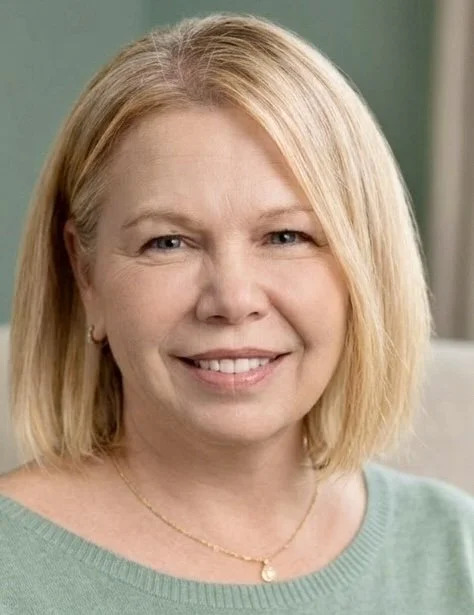 A smiling woman with blonde hair, wearing a light green top and a gold necklace, indoors with a green wall in the background.