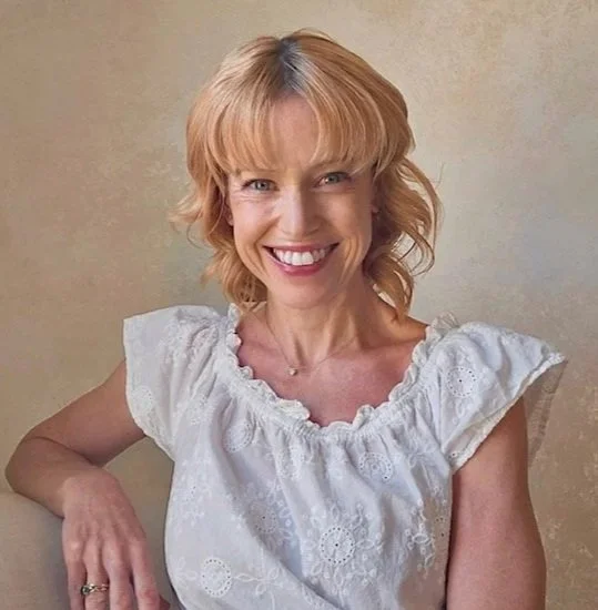 A woman with shoulder-length blonde hair wearing a white eyelet blouse, smiling and sitting on a beige couch against a plain beige wall.
