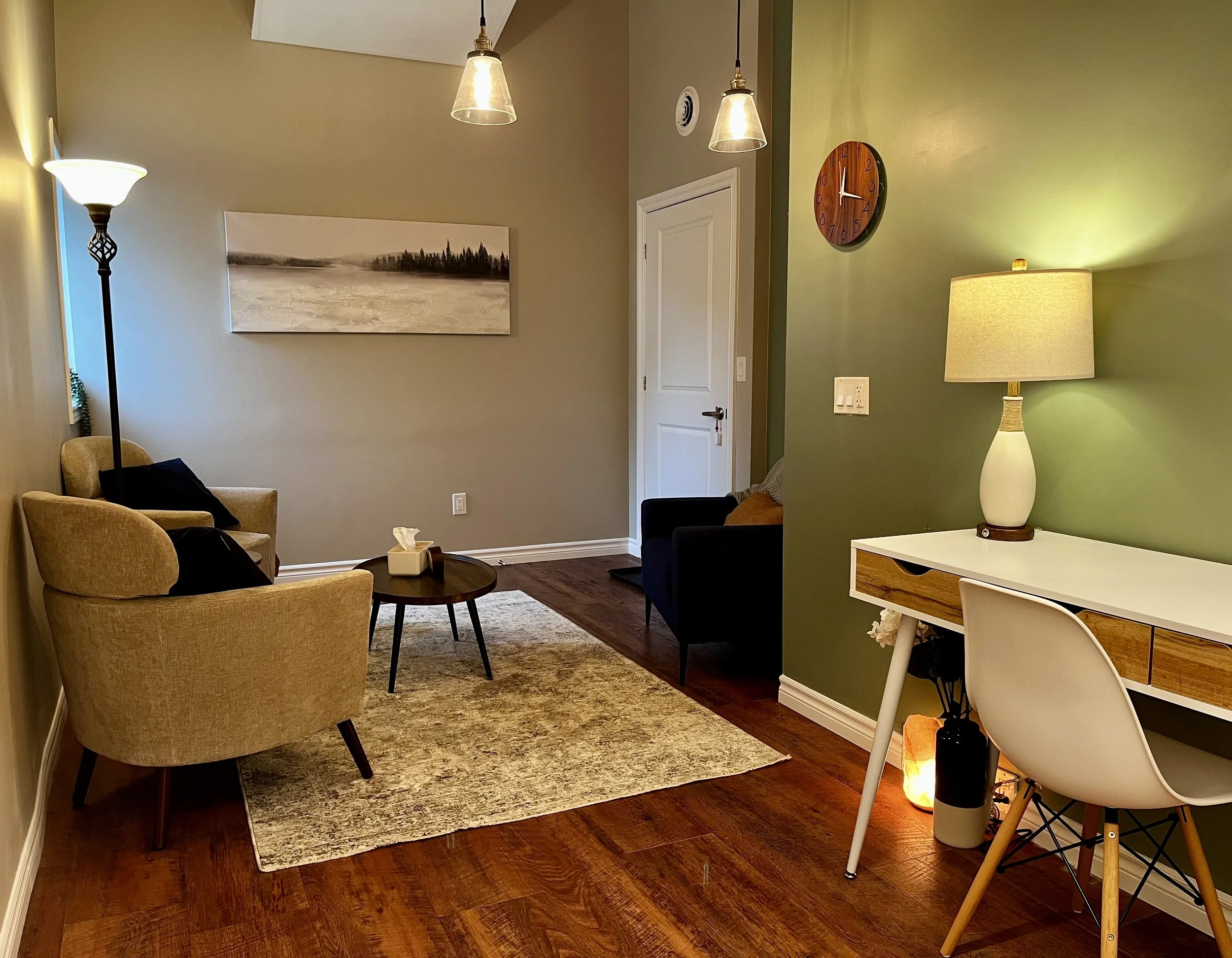 Cozy living room with green walls, featuring armchairs, coffee table, floor lamp, wall art, desk with chair, table lamp, and wooden flooring.