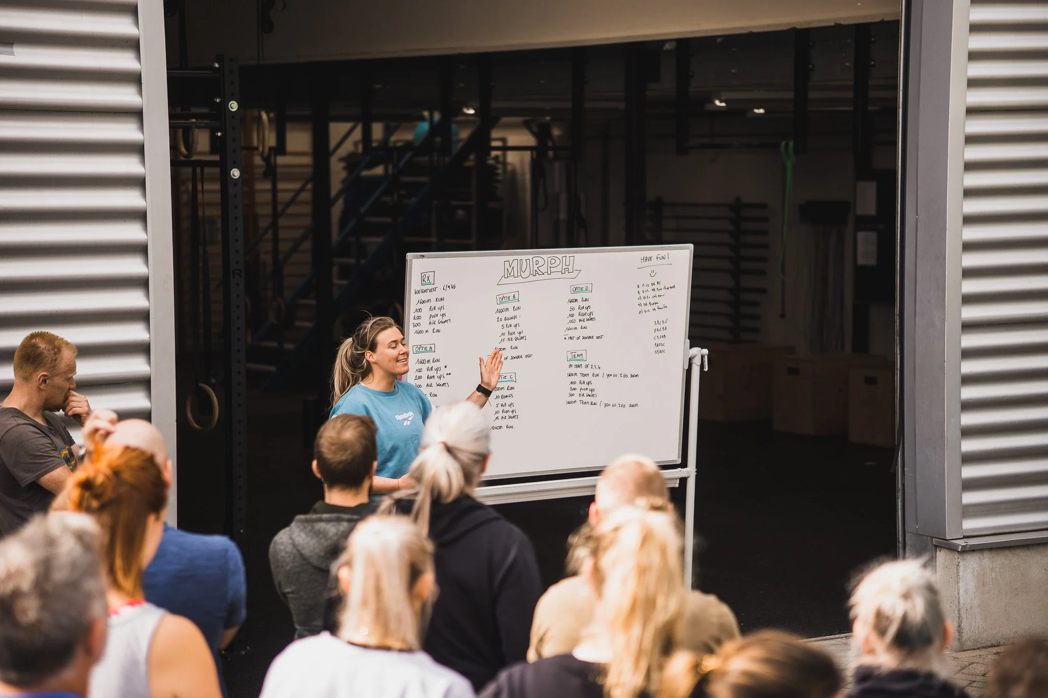 Coach Kim van Black Oak in een lichtblauw shirt staat voor een wit bord en legt een workout uit aan een luisterende groep mensen in een sportschool.