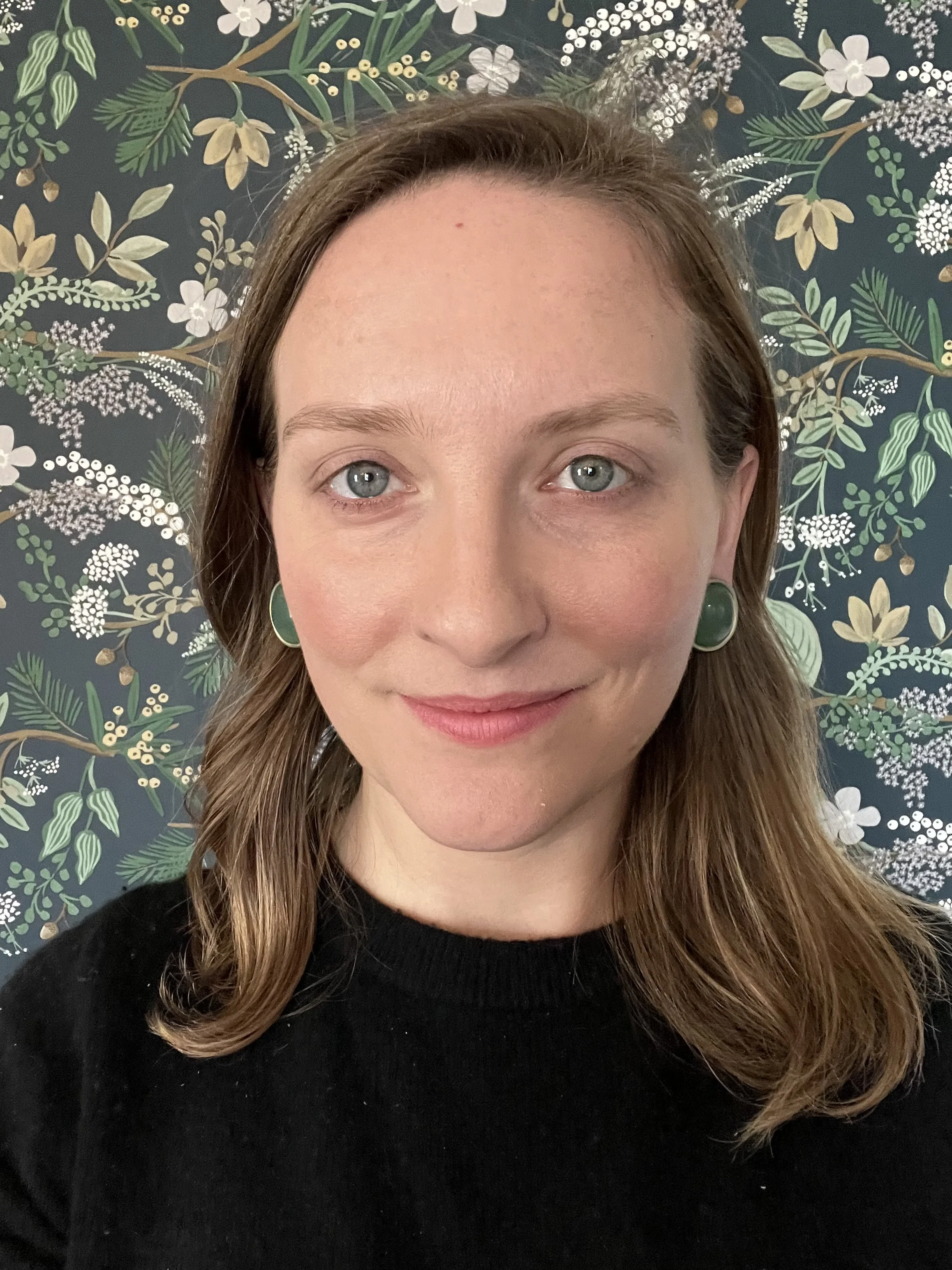 Blond woman smiles into camera. She has shoulder length hair and is wearing a black shirt. There is a green floral wall behind her.