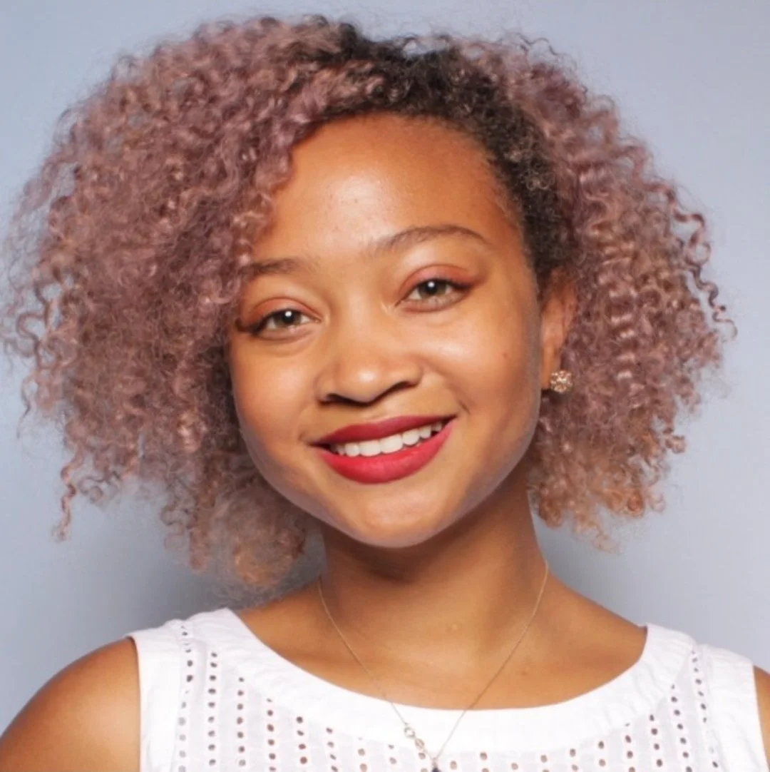 A woman with curly hair is wearing a white tank top and smiling into the camera