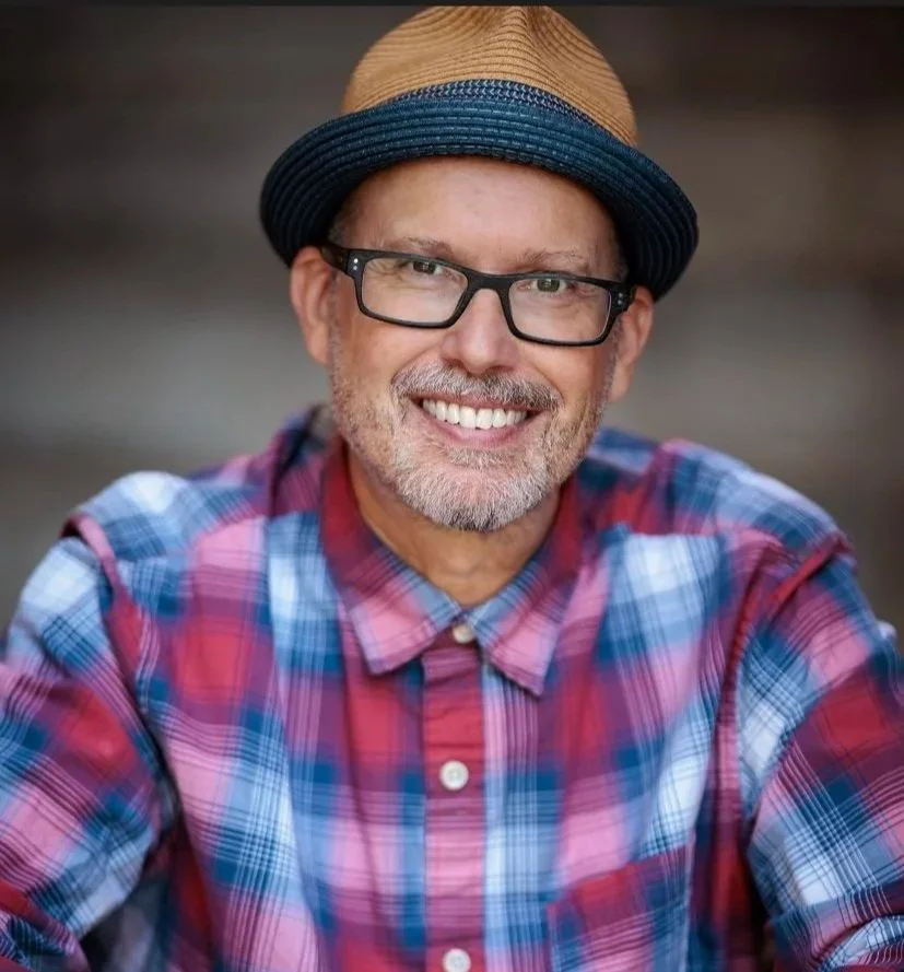 A man smiles into the camera. He is wearing a straw hat, black glasses, and a red and blue plaid shirt.