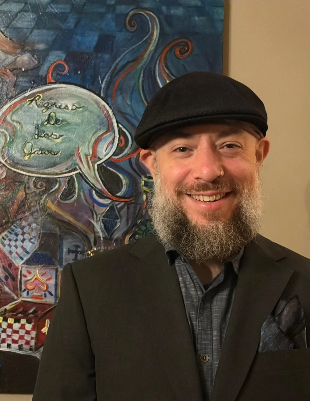 A bearded man wearing a hat stands in front of a painting and smiles