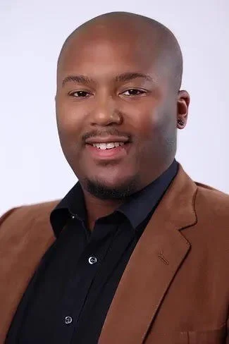 A young man smiles at the camera. He is wearing a black shirt and brown suit jacket.