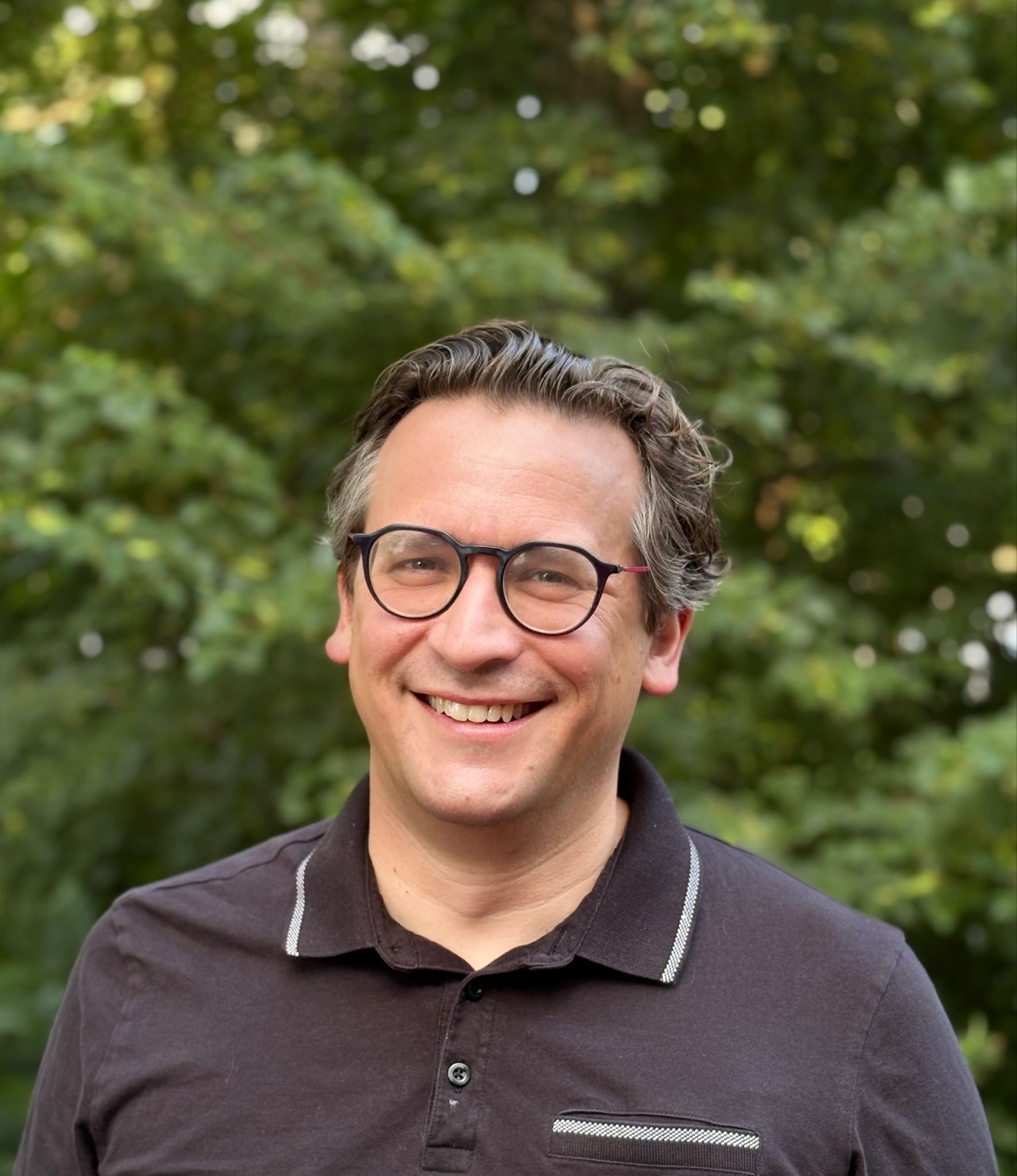 A man with black rimmed glasses and dark hair stands in front of trees, smiling