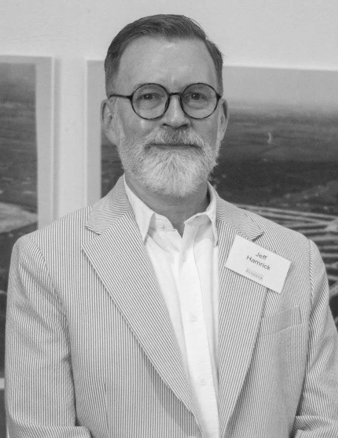 Black and white photo of a smiling man with short hair and a neatly trimmed beard. He is wearing glasses and a white shirt with a suit jacket.