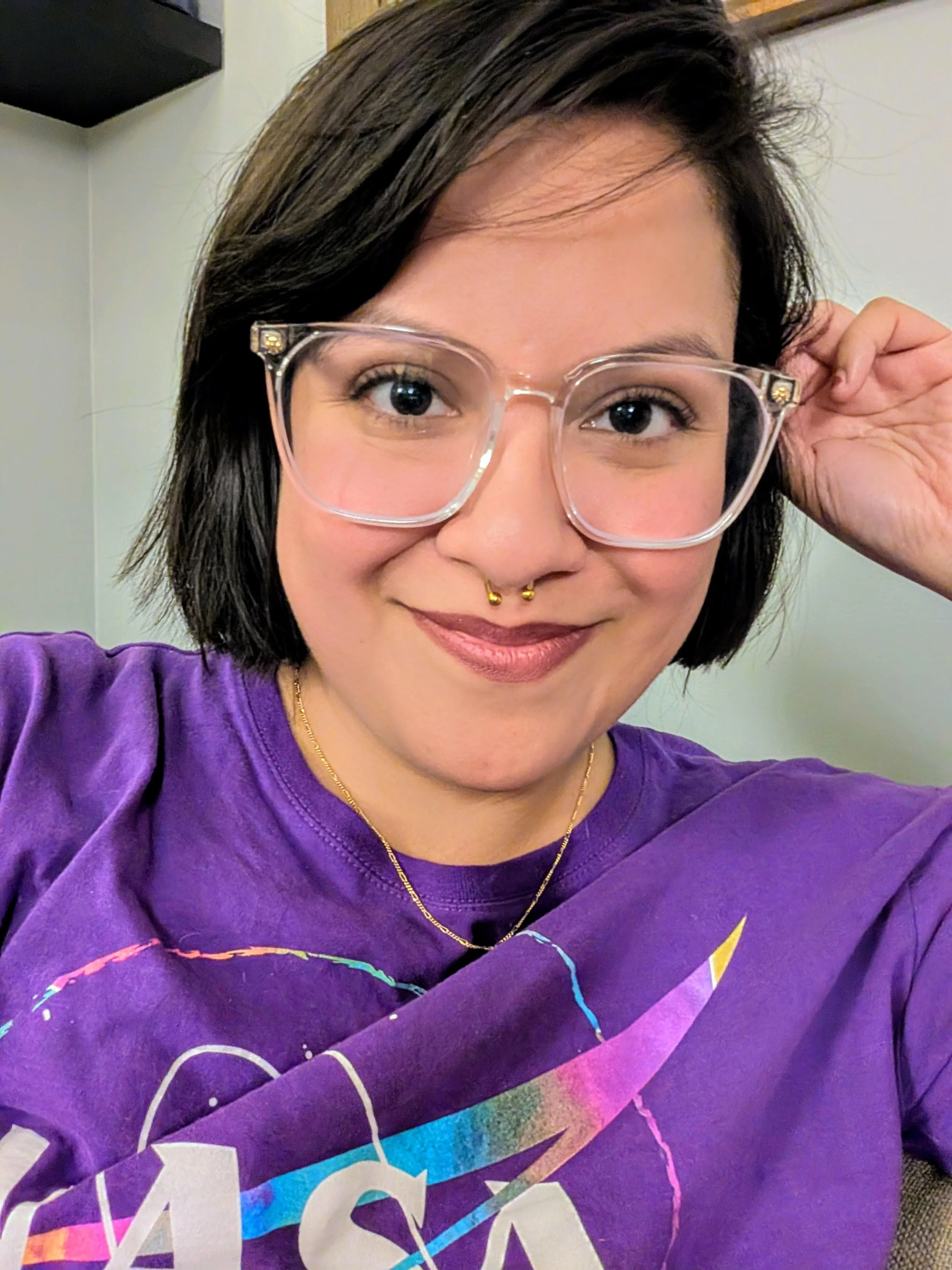 A woman with chin length, dark hair and glasses smiles at the camera. She is wearing a purple NASA shirt.