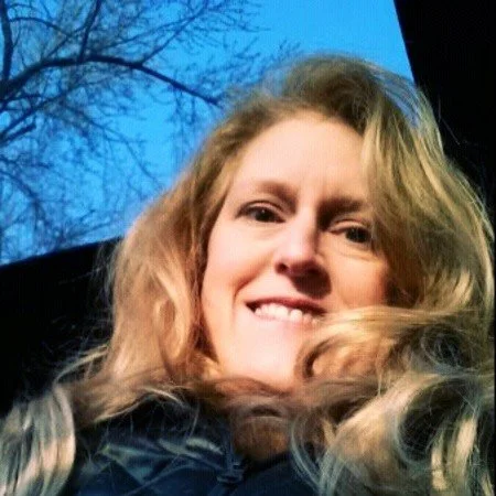 A blond woman looks down into the camera. She it outside that the sky and tree branches are visible above her
