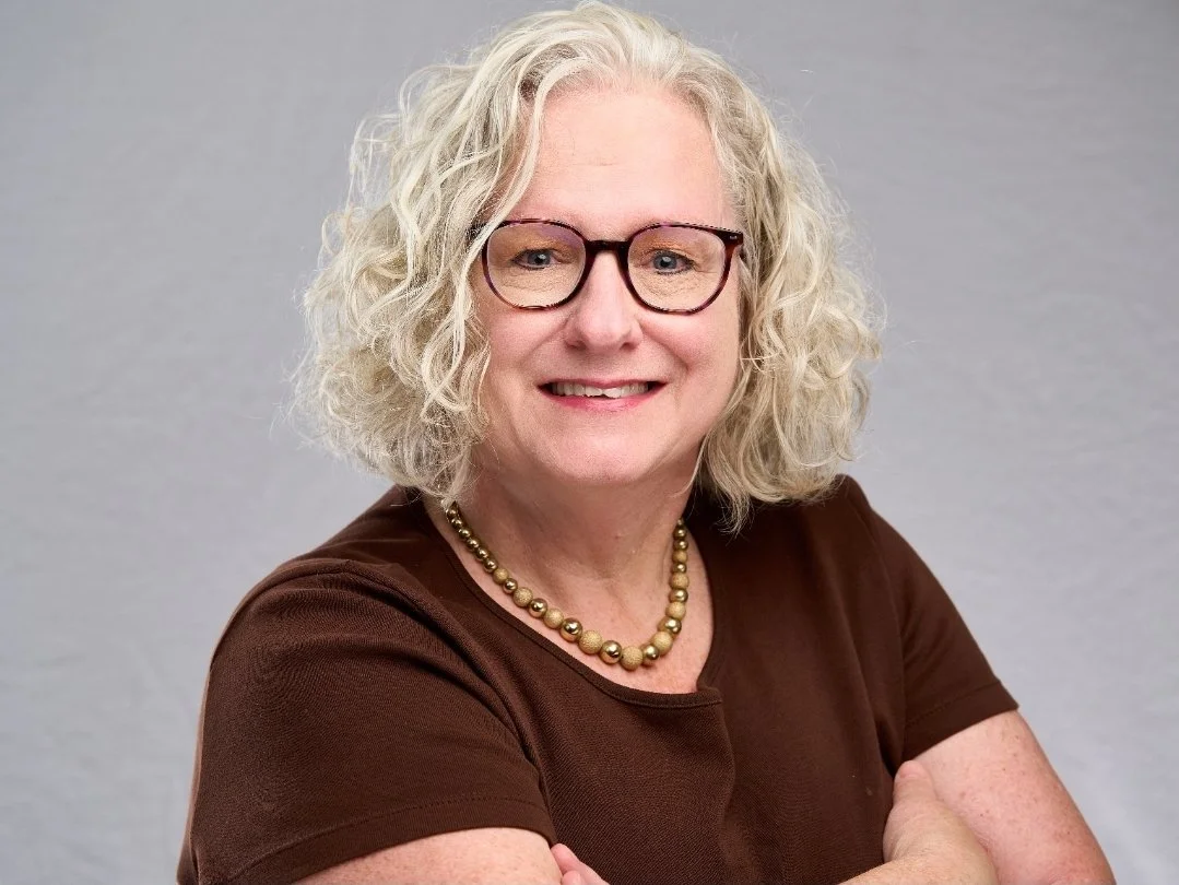 A woman with curly blonde hair smiles into the camera, arms crossed