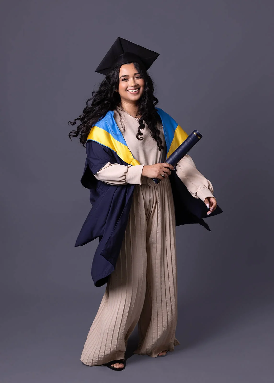 Woman in graduation gown and cap holding a diploma, standing against a gray background.