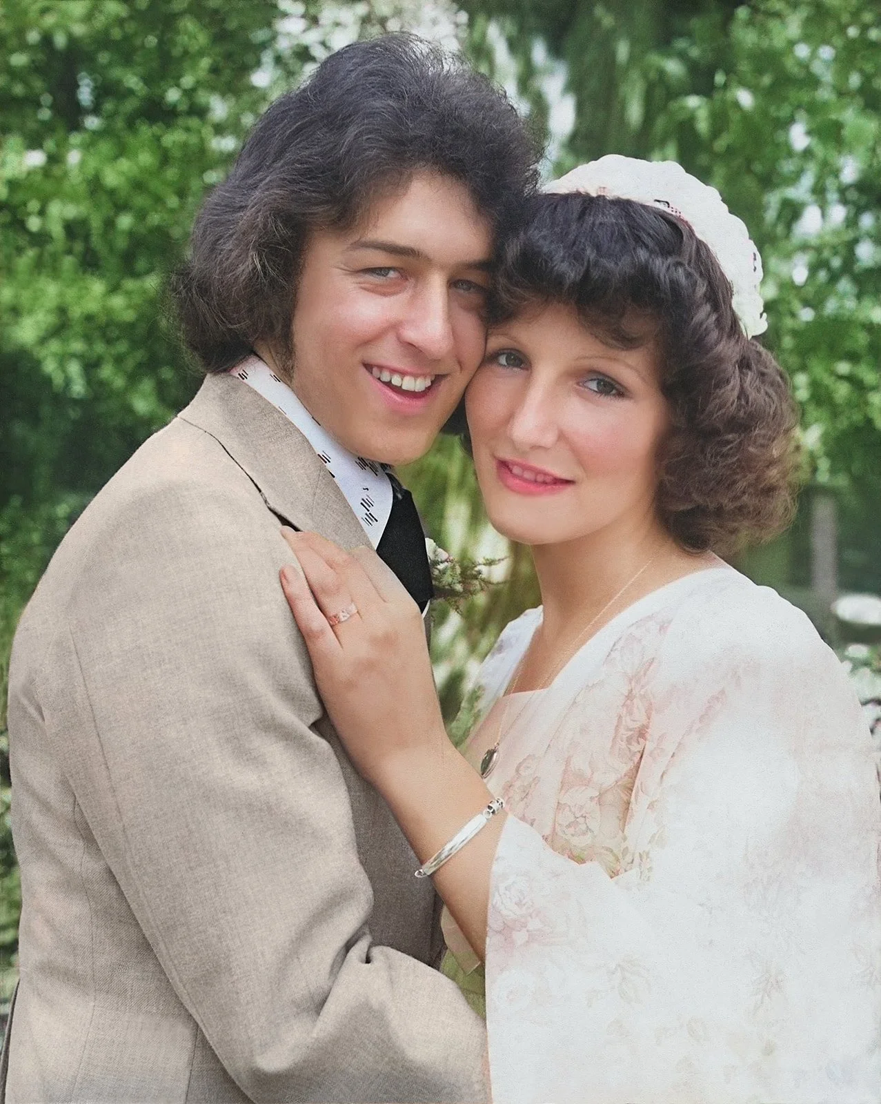 A young man and woman are closely embracing outdoors with green trees in the background. They are smiling and appear happy.