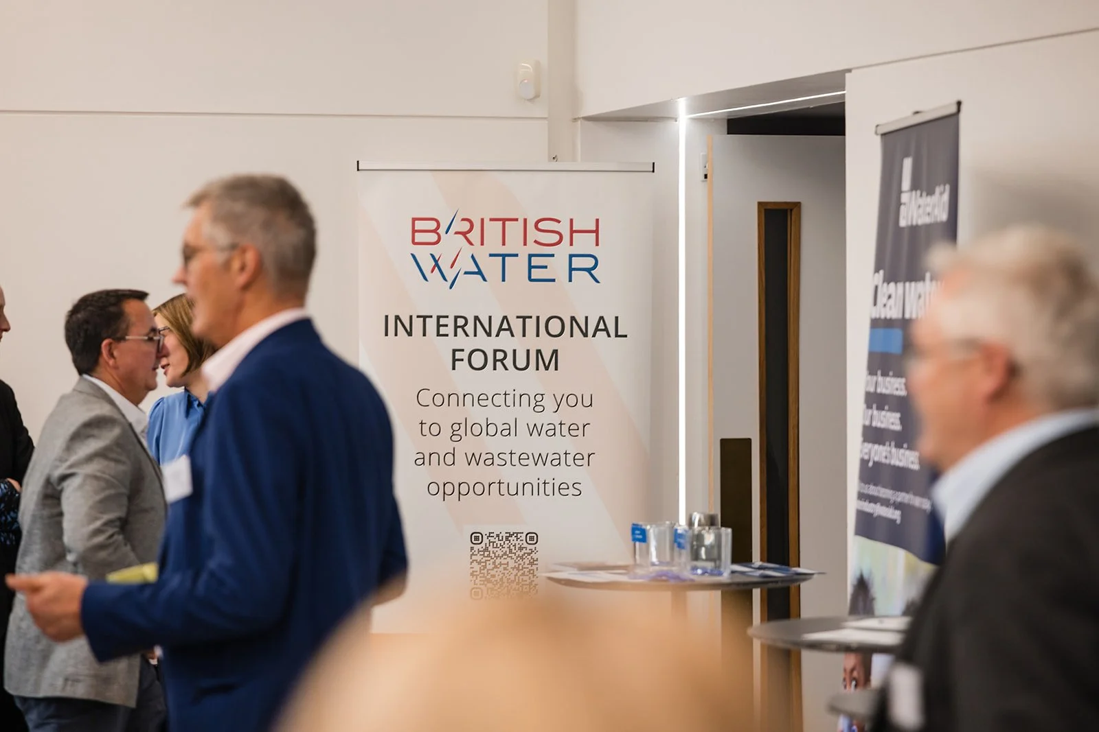 Attendees at the British Water International Forum event, engaging in conversation. A large banner displaying the event name and mission statement about connecting to global water and wastewater opportunities is visible.