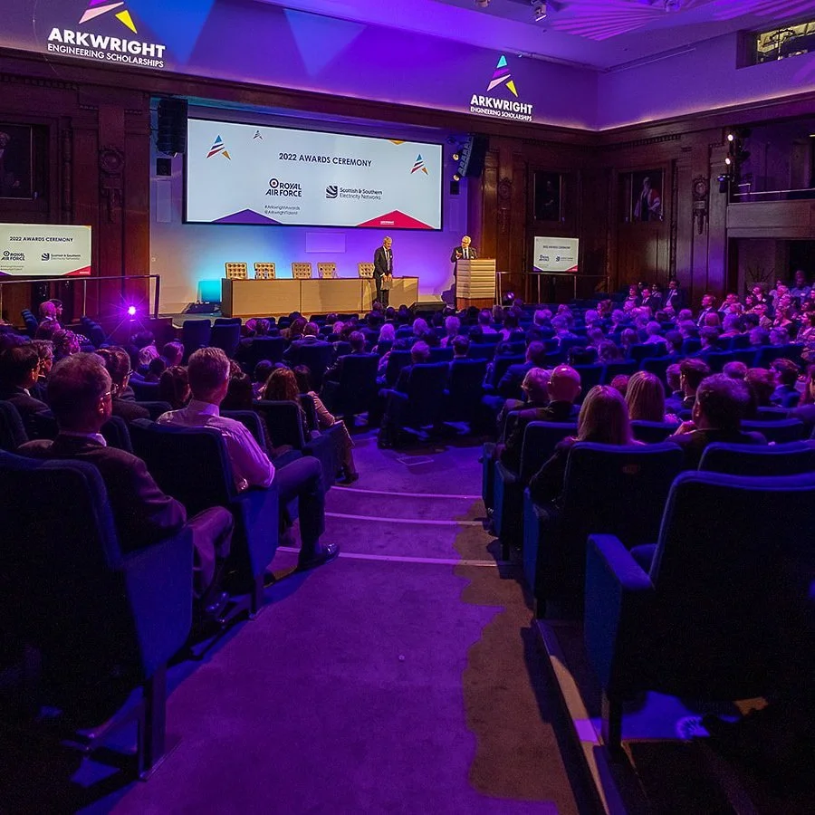 People attending an awards ceremony in a large auditorium with a stage and a large screen displaying the event's details. The event is titled '2022 Awards Ceremony' organized by Arkwright Engineering Scholarships, Royal Air Force, and Scottish & Southern Electricity Networks.