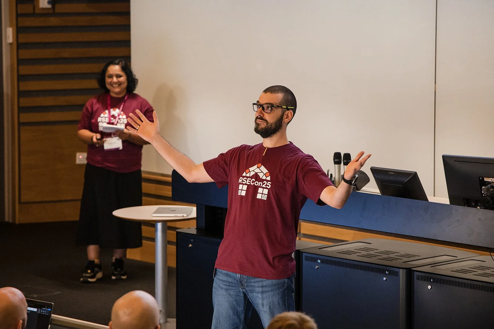 A man wearing glasses and a maroon T-shirt with 'RSEcon25' logo is giving a presentation in a conference room, with a woman smiling and standing in the background.
