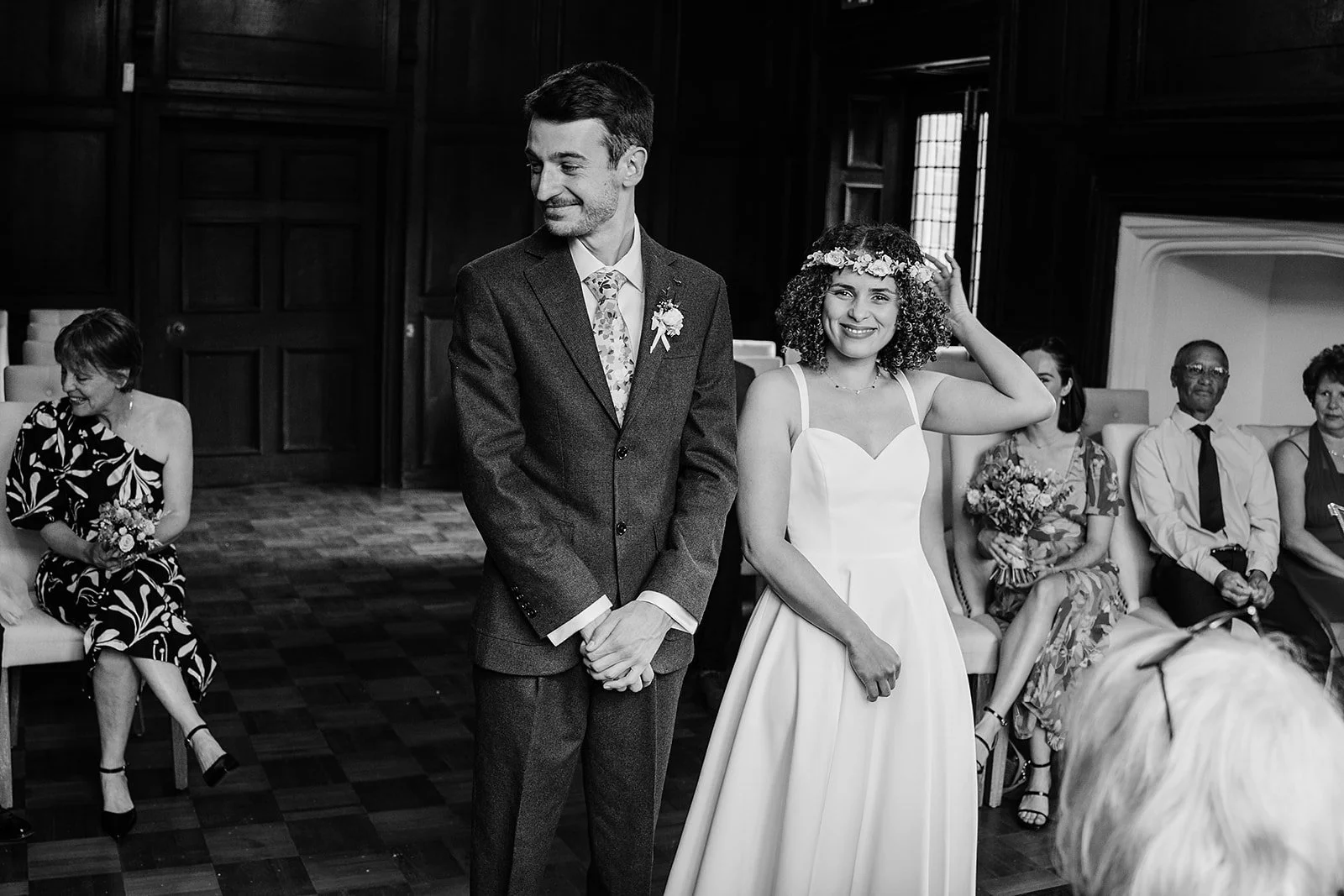 A black and white photograph of a bride and groom standing in a room during a wedding ceremony. The bride is smiling and touching her floral crown, while the groom stands with his hands clasped, smiling softly. They are surrounded by seated guests, some holding flowers, inside a wood-paneled room with large windows.