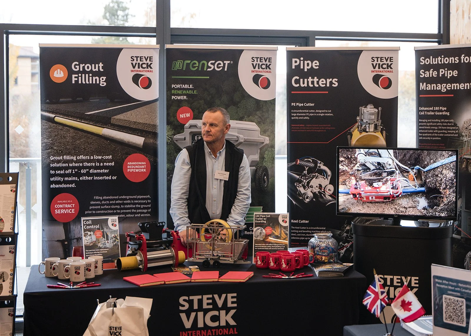 A man standing behind a table at an exhibition booth with posters and products related to pipe and conduit installation and management. The booth displays banners with text about grout filling, trenchless pipe installation, pipe cutters, and safe pip