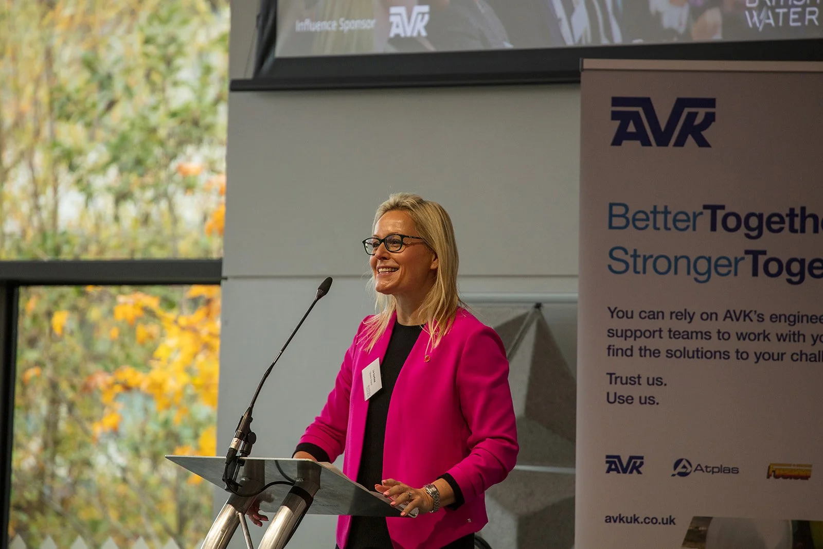 A woman with blonde hair and glasses wearing a pink blazer standing behind a podium, smiling during a speech at an indoor event with large windows showing trees with autumn leaves.
