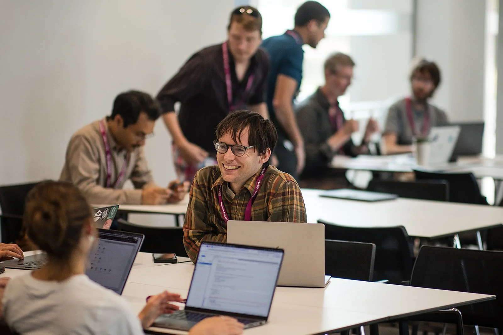 People working and collaborating in a modern office meeting room, some seated at laptops and others standing and discussing.