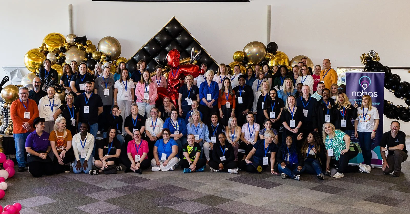 Group photo of diverse people at a conference or event, with balloons and decorations in the background, and a NABAS banner on the right.