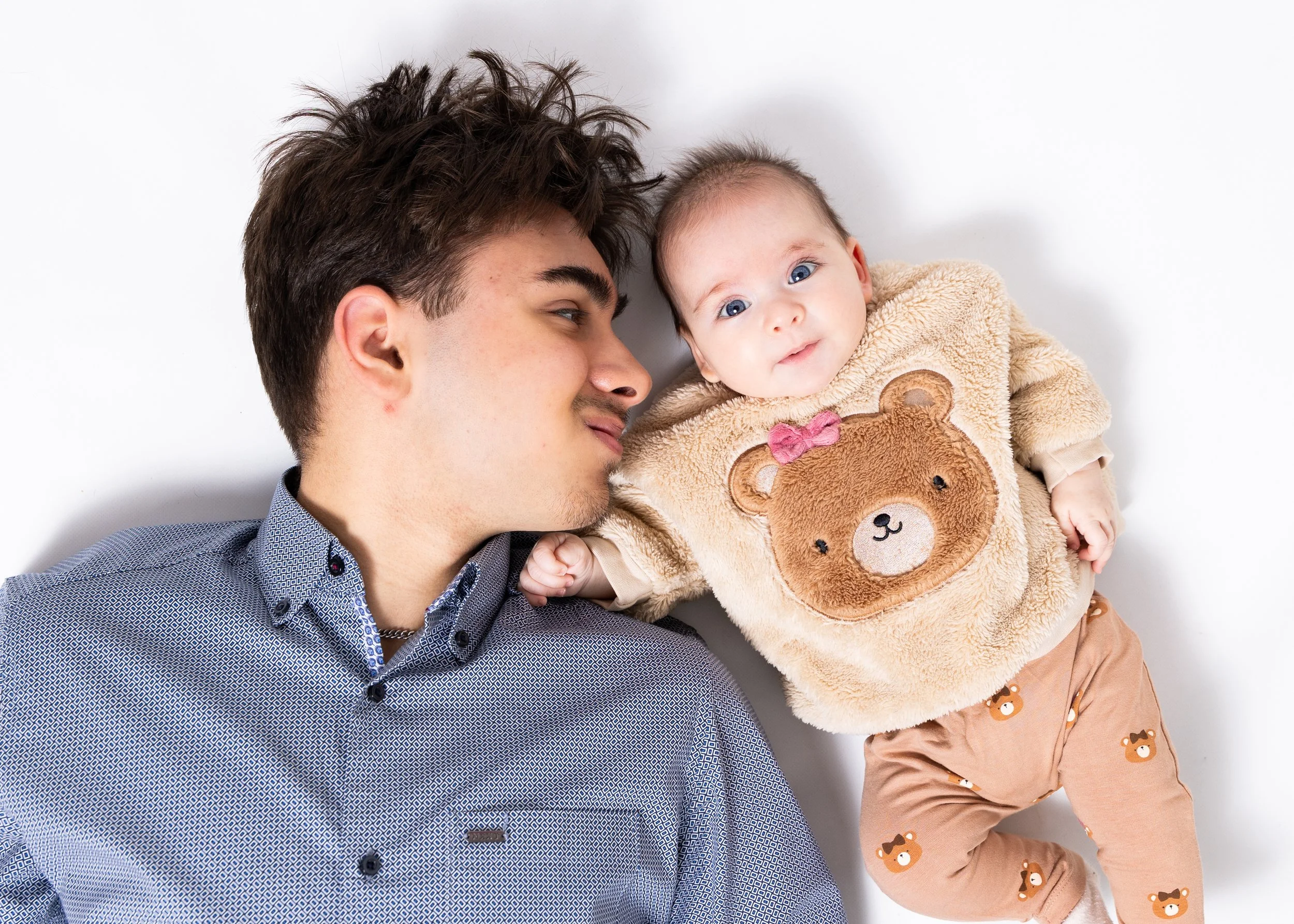 A young man lying on his side next to a baby girl on a white background, both smiling and looking at the camera. The baby is wearing a teddy bear sweatshirt and matching pants.
