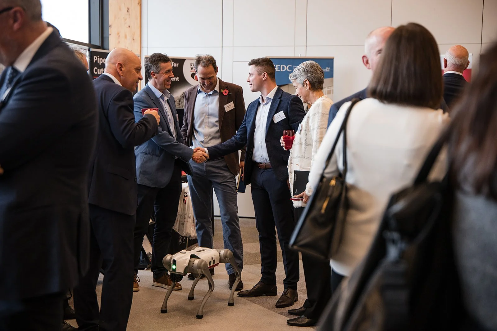 Business professionals at a networking event, shaking hands and talking, with a small robotic dog on the floor.