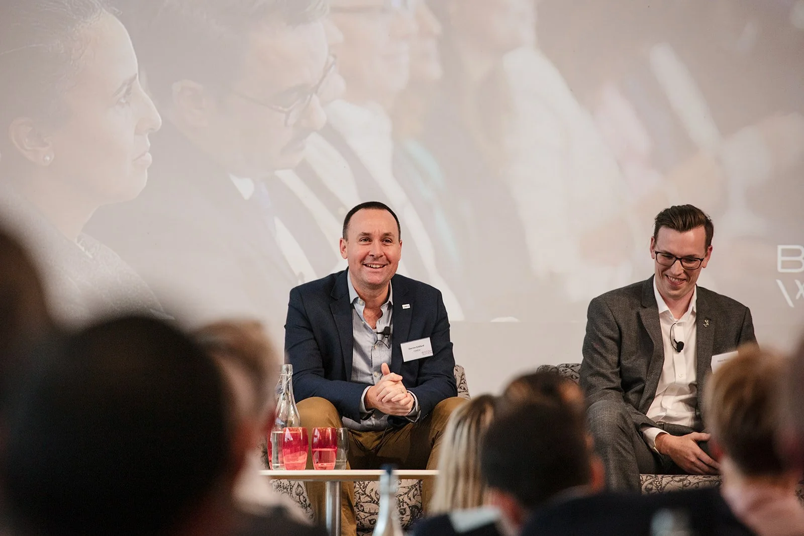 Two men sitting on a stage during a panel discussion, with an audience in front of them. The man on the left is smiling, wearing a dark blazer, light shirt, and tan pants. The man on the right is also smiling, wearing a gray checked suit and glasses.