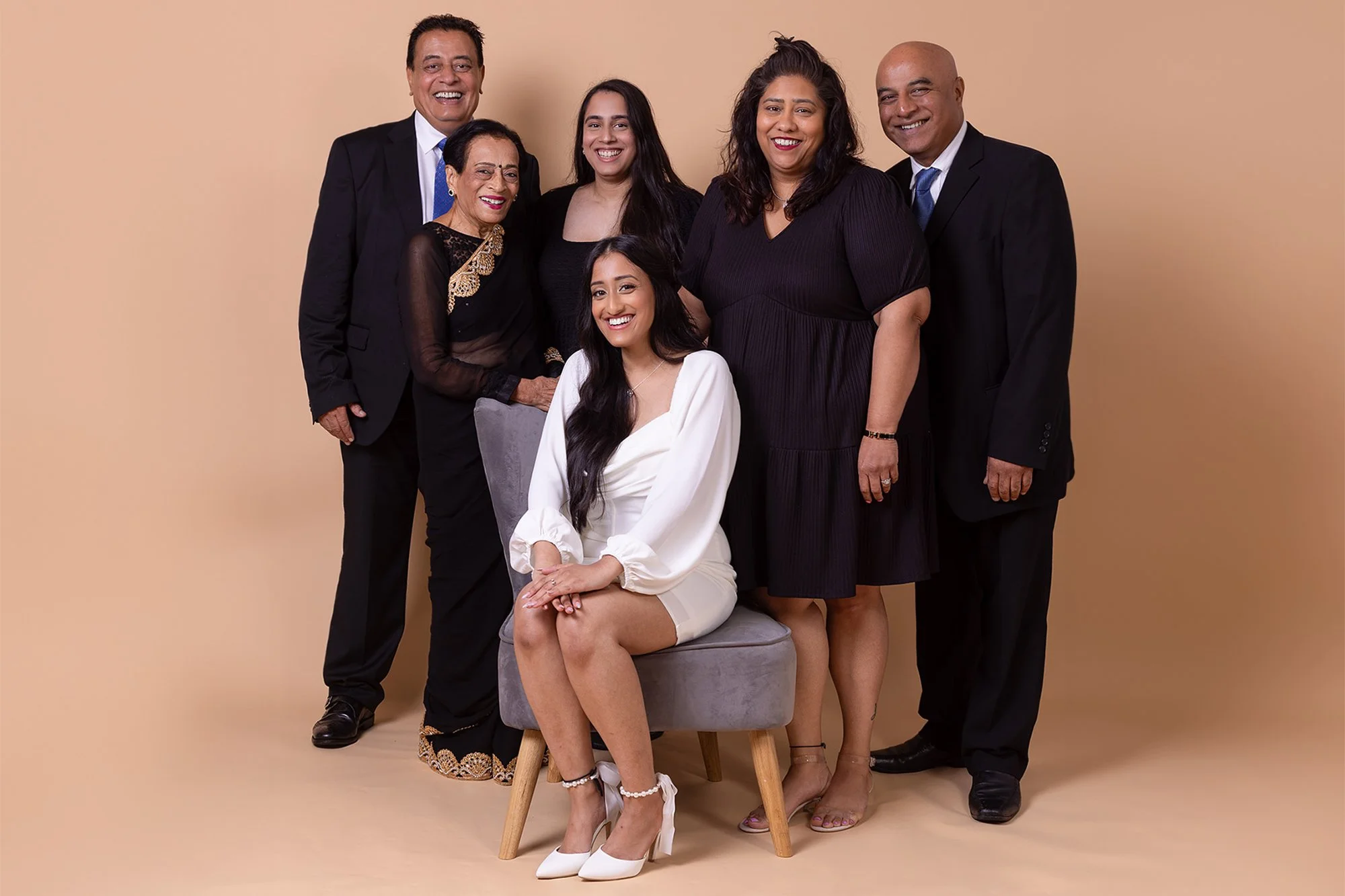 A group of six ethnically diverse people posing together, dressed in formal attire, against a beige background. One woman is seated on a chair, wearing a white dress and high heels, while the others stand around her and smile at the camera.