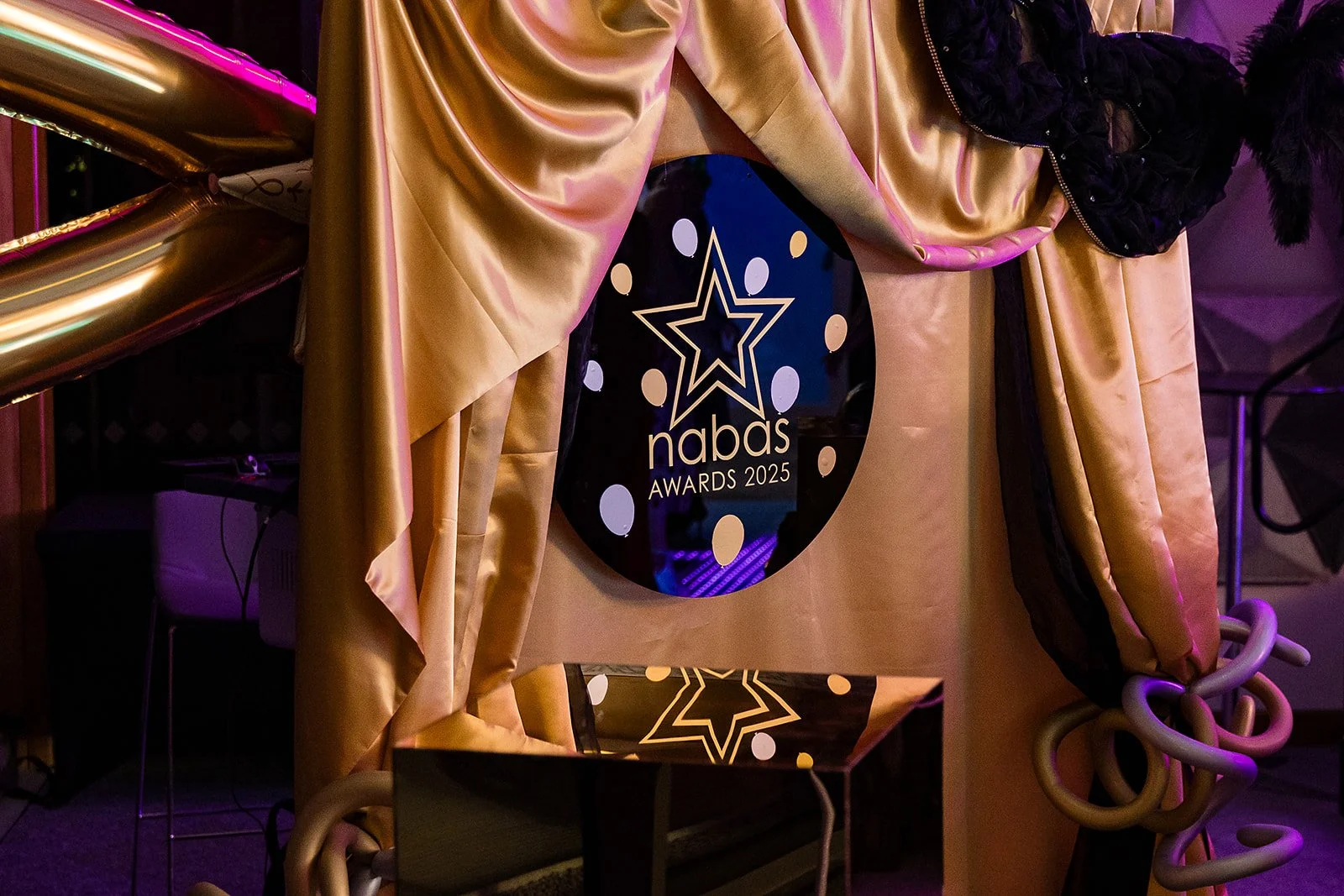 A reflective black circular sign with a star logo and text that reads 'nabas Awards 2025,' placed on a table with shiny beige and black curtains and purple lighting in the background.