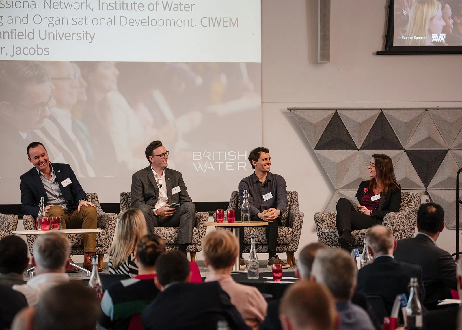 Four people sitting on stage participating in a panel discussion at a conference. Audience members seated in front watching. Large screen behind showing text and a image of a previous panel, with the words 'British Water' visible.