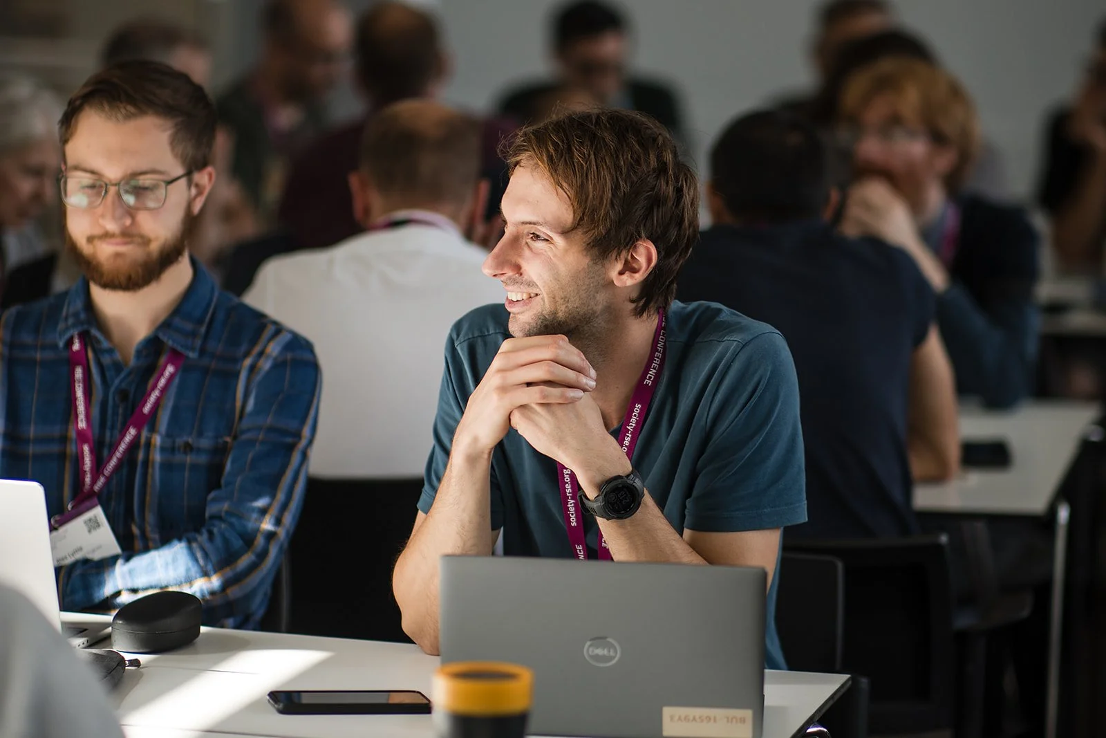 A group of young adults attending a conference or workshop, sitting at tables with laptops and engaged in discussions, with some smiling and others focused.