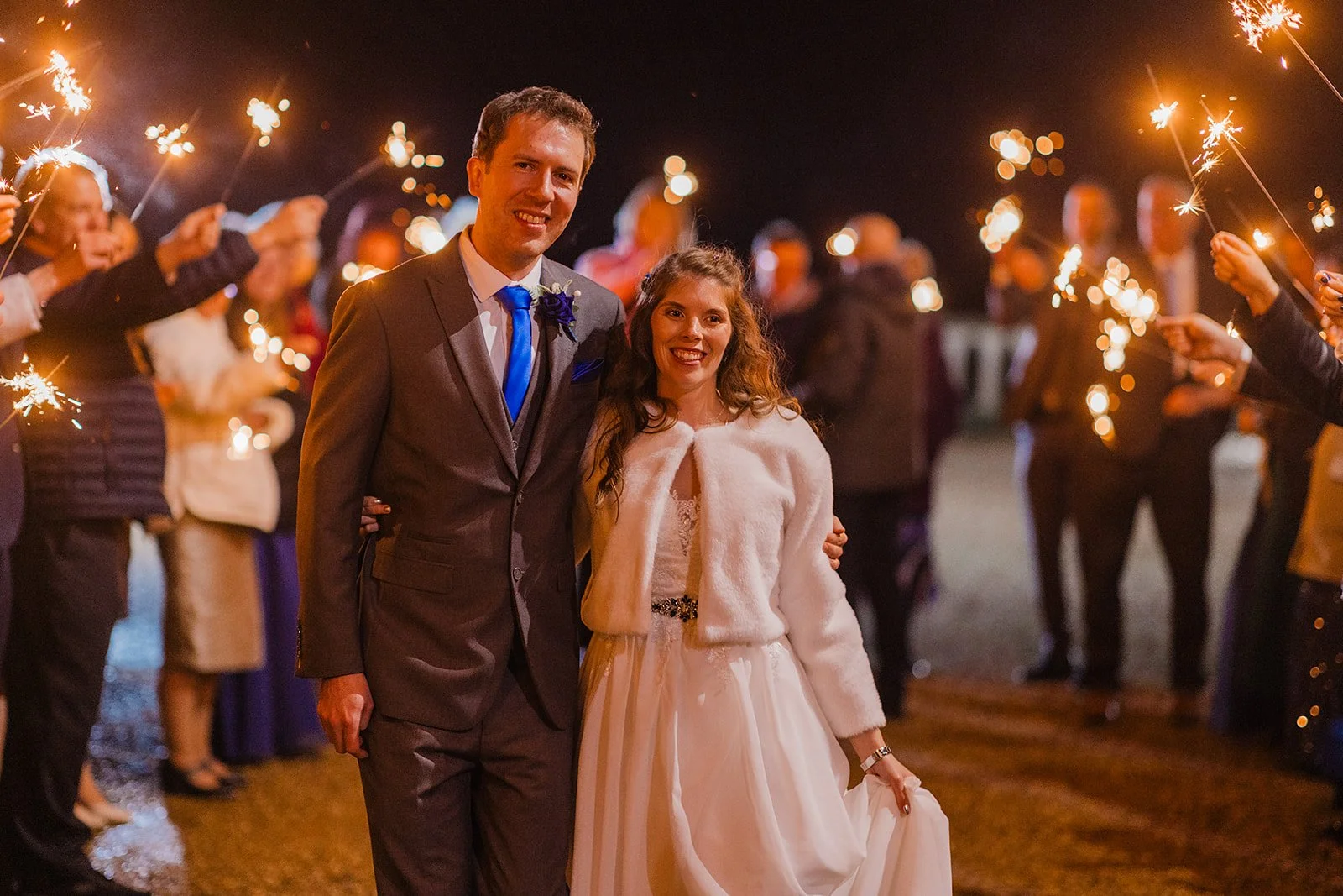 A bride and groom walking outside at night, surrounded by guests holding sparklers in celebration.