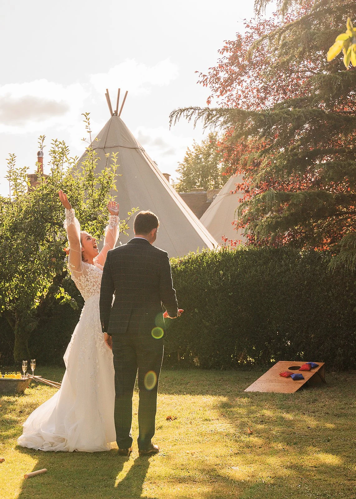 A bride and groom standing outdoors during a wedding ceremony in the late afternoon sunlight, with tipi tents and trees in the background.
