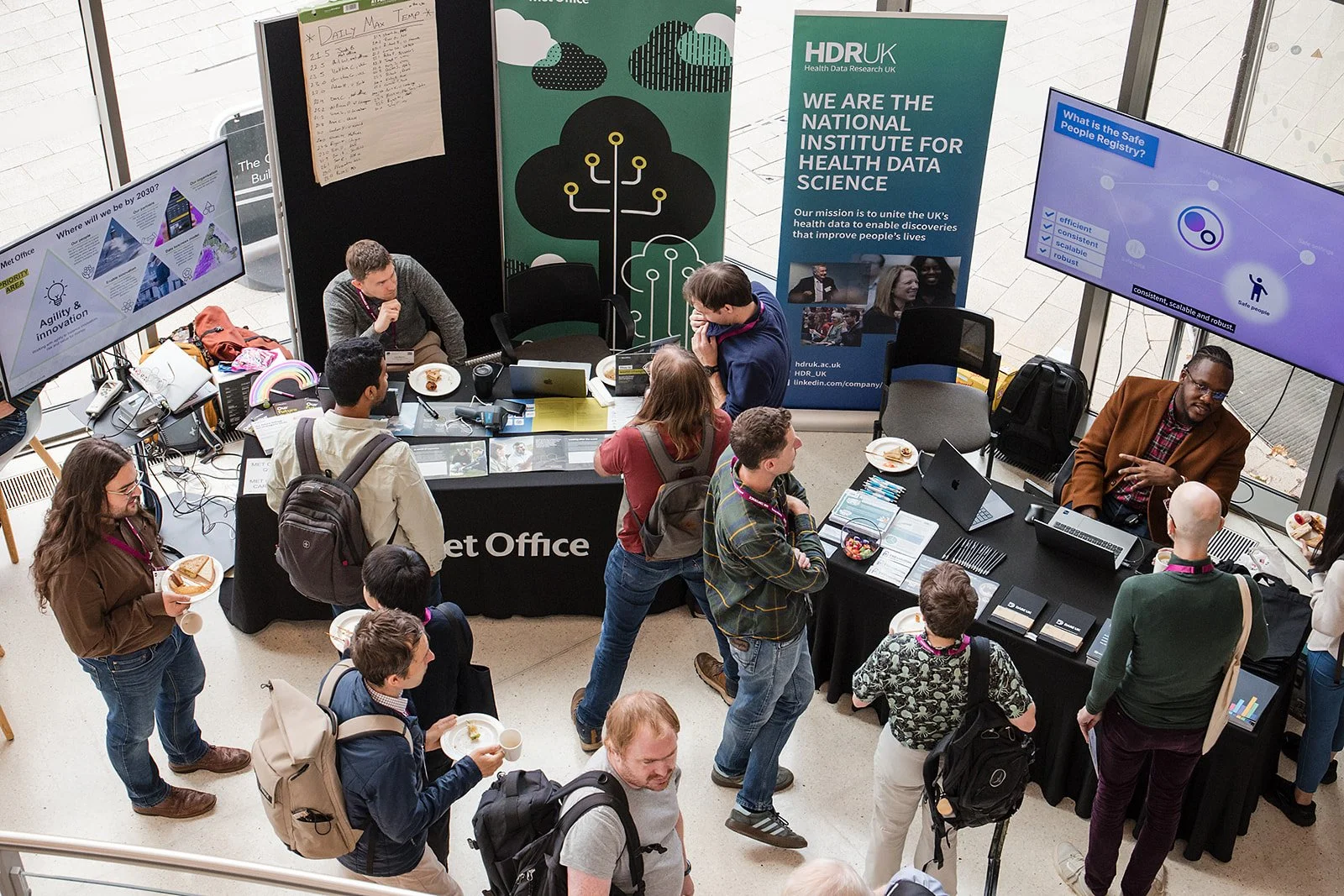 At a conference booth, attendees are gathered around two large screens displaying information about health data research and the safe people registry, with posters and laptops on the tables.