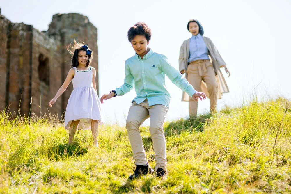 Three children playing on a grassy hillside with an old building in the background.